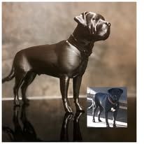 Een donkermetalen sculptuur van een hond die op een reflecterend oppervlak staat, naast een kleine foto van een zwarte hond.