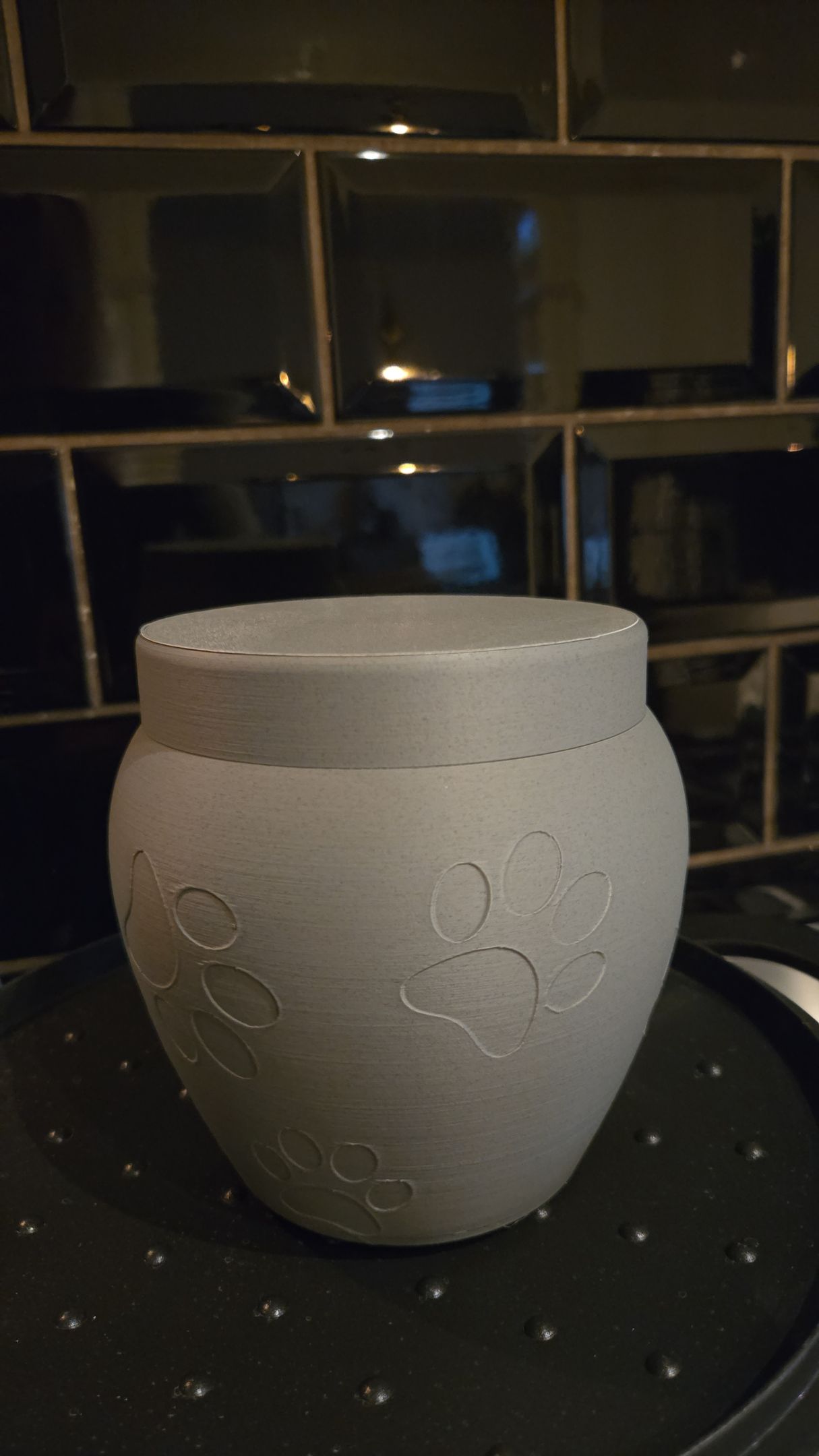 Een grijze, matte keramische urn voor de crematie van een huisdier, met een subtiel reliëf van pootafdrukken, op een fornuis.