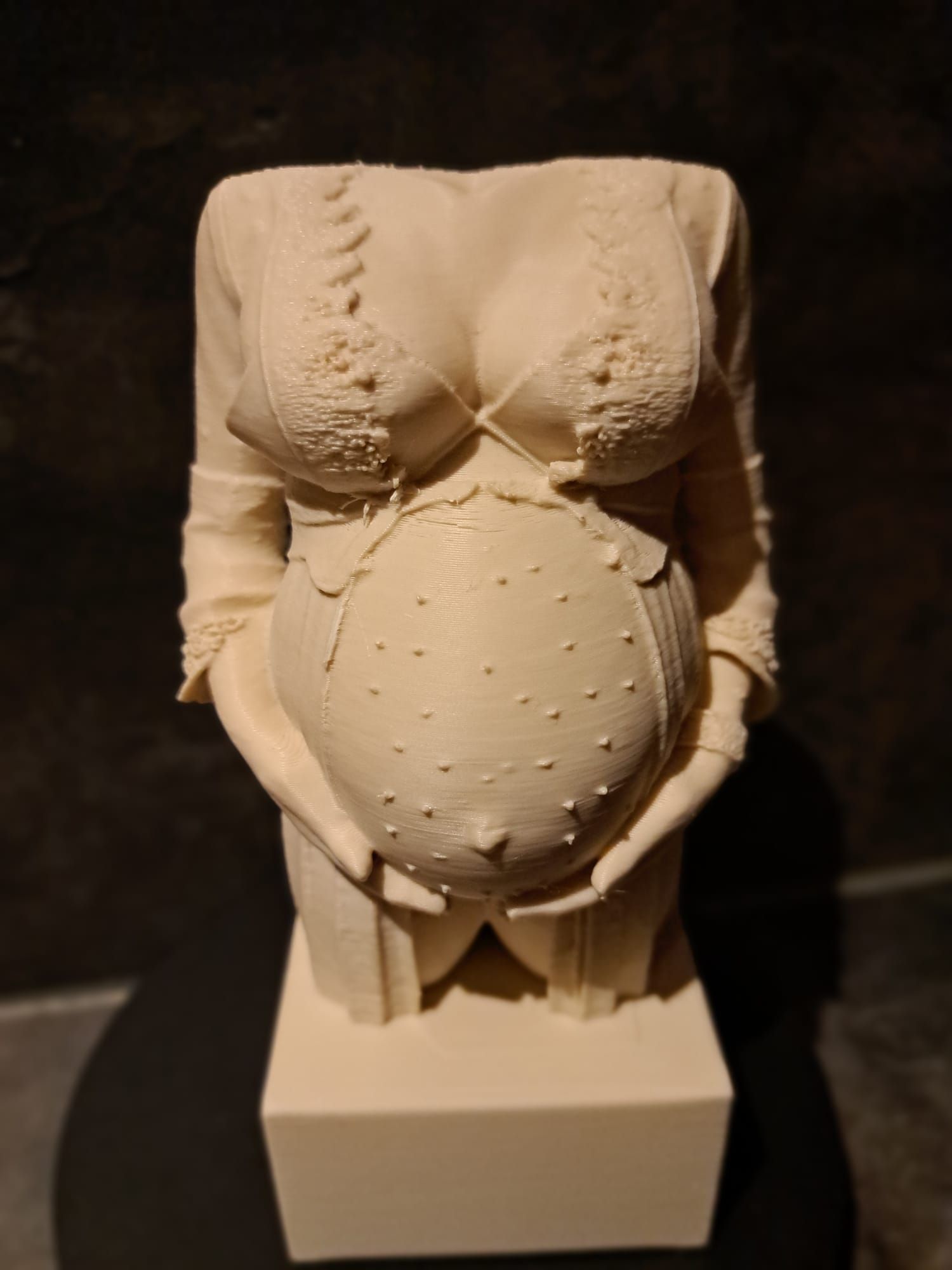 Een lichtgekleurd beeld van een zwangere torso, tot aan de taille, met kanten kleding aan, rustend op een sokkel.