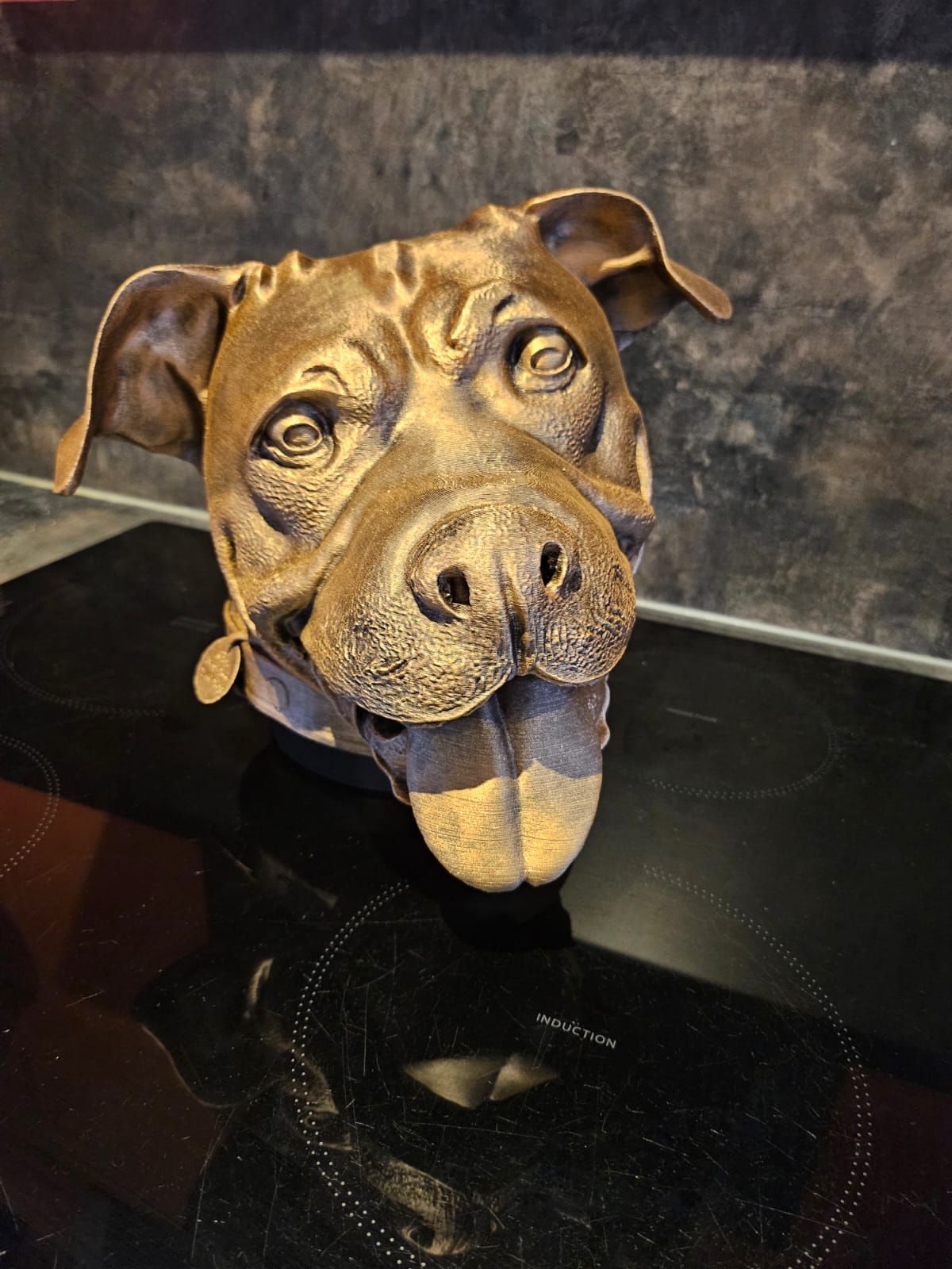 Een bronzen sculptuur van een pitbullkop met een vrolijke uitdrukking en uitgestoken tong, zittend op een fornuis.