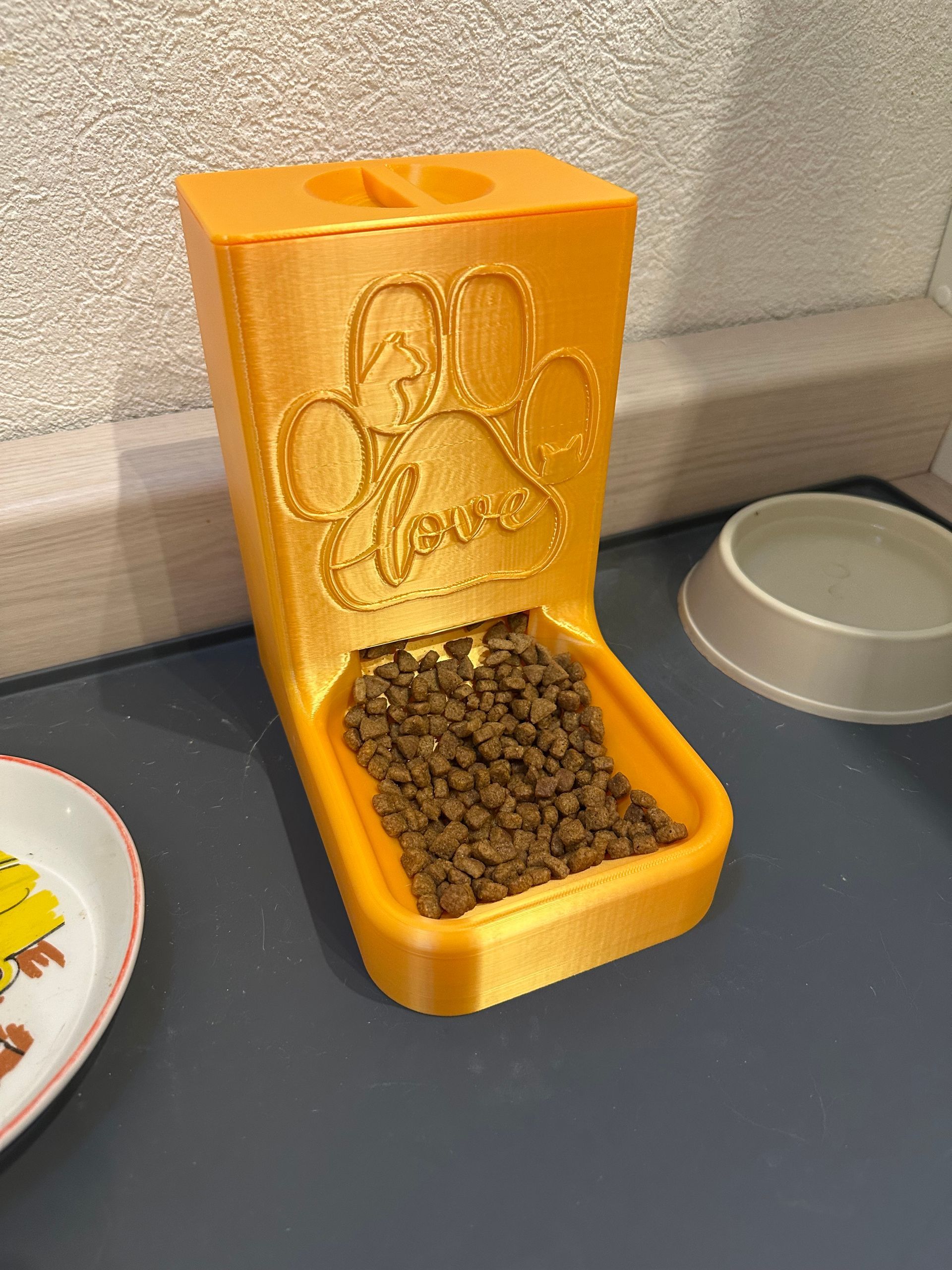 Een gele, 3D-geprinte voerdispenser voor huisdieren met een pootafdruk en het woord 