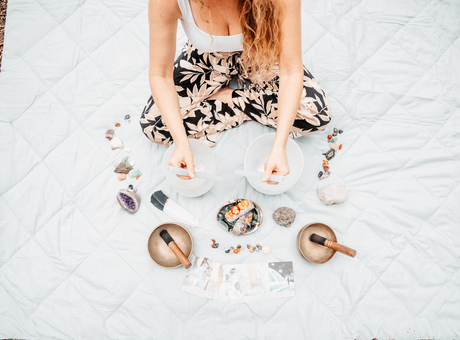 a woman sits on a blanket playing sound healing bowls surrounded by oracle cards, and tibetan singing bowls