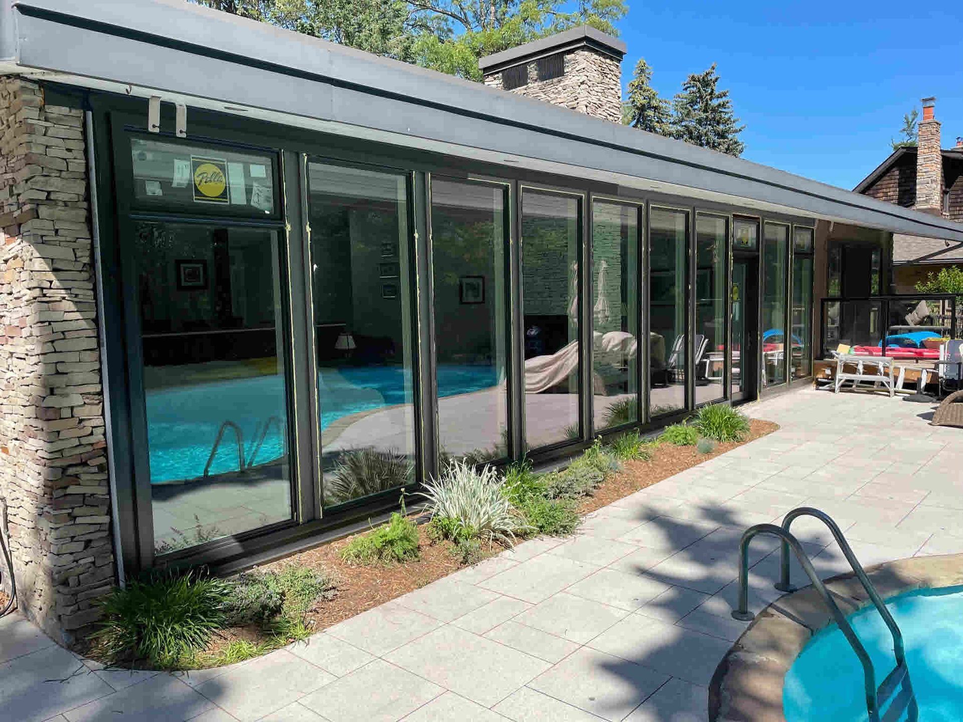 Building with large windows next to a pool. Stone wall, patio, and blue pool water are visible.
