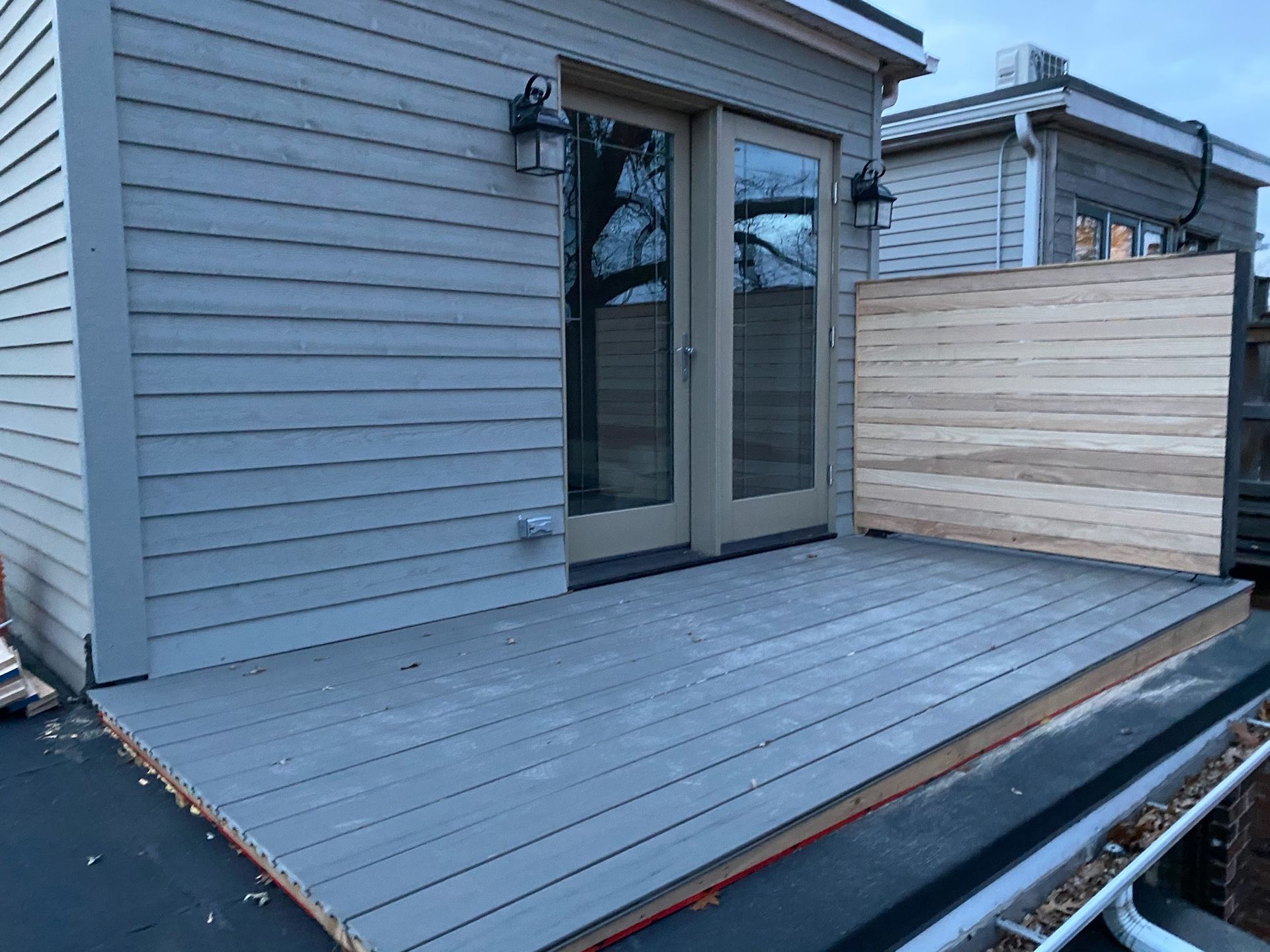 A rooftop deck with gray composite decking, double doors, and a wood privacy screen.