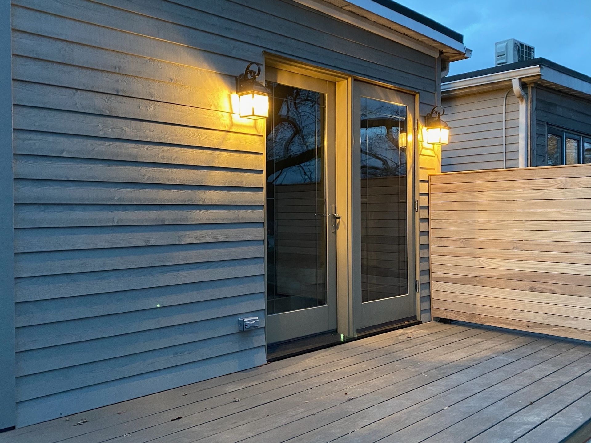 Gray siding with two glass doors, two lights, and a wooden deck.