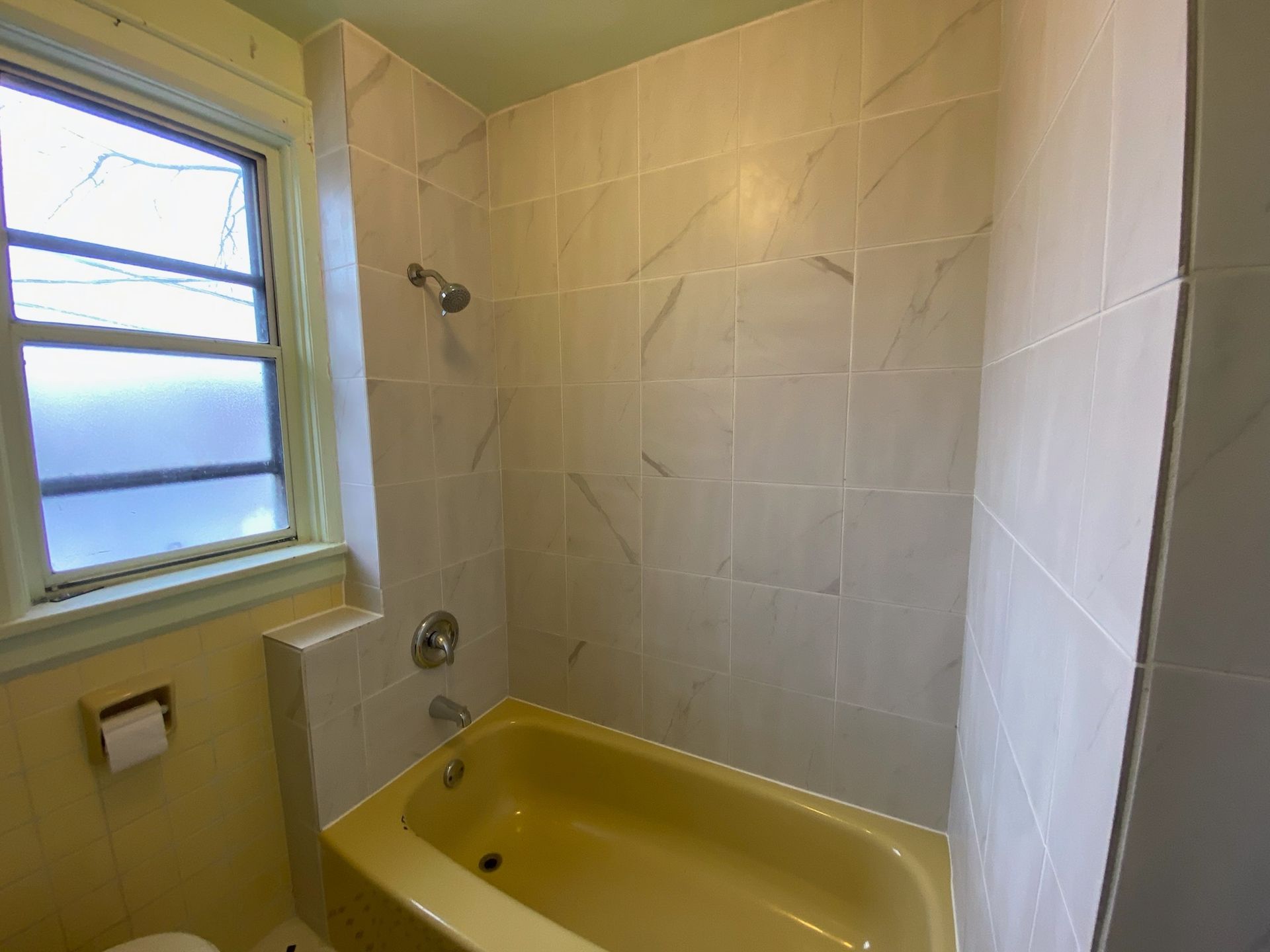 Bathroom with yellow tub, gray tile walls, window, and shower fixtures.