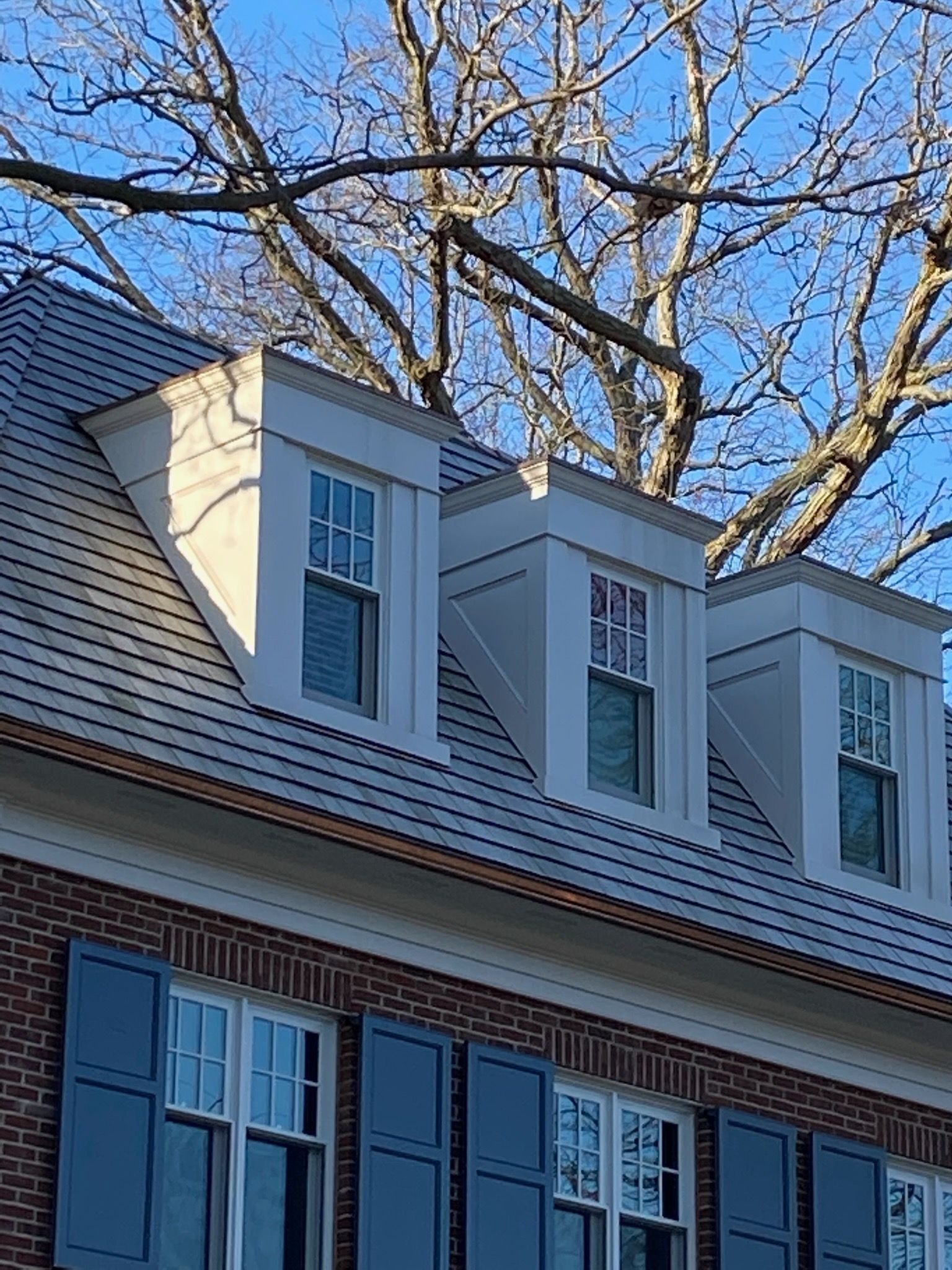Brick house with dormers, blue shutters, and bare tree branches against a blue sky.