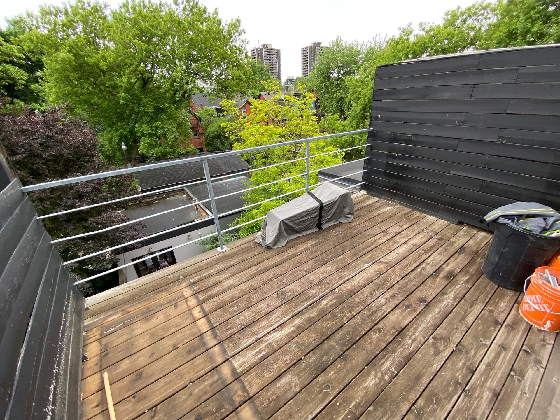 Rooftop deck with wooden planks, metal railing, and black paneling, overlooking trees and buildings.