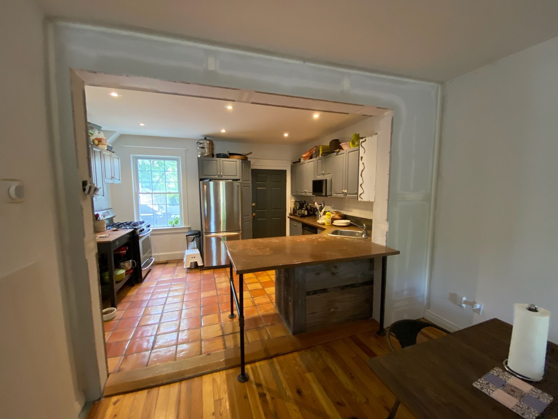 Kitchen with doorway opening into a room. Features a wooden table with metal legs and terracotta tile floor.