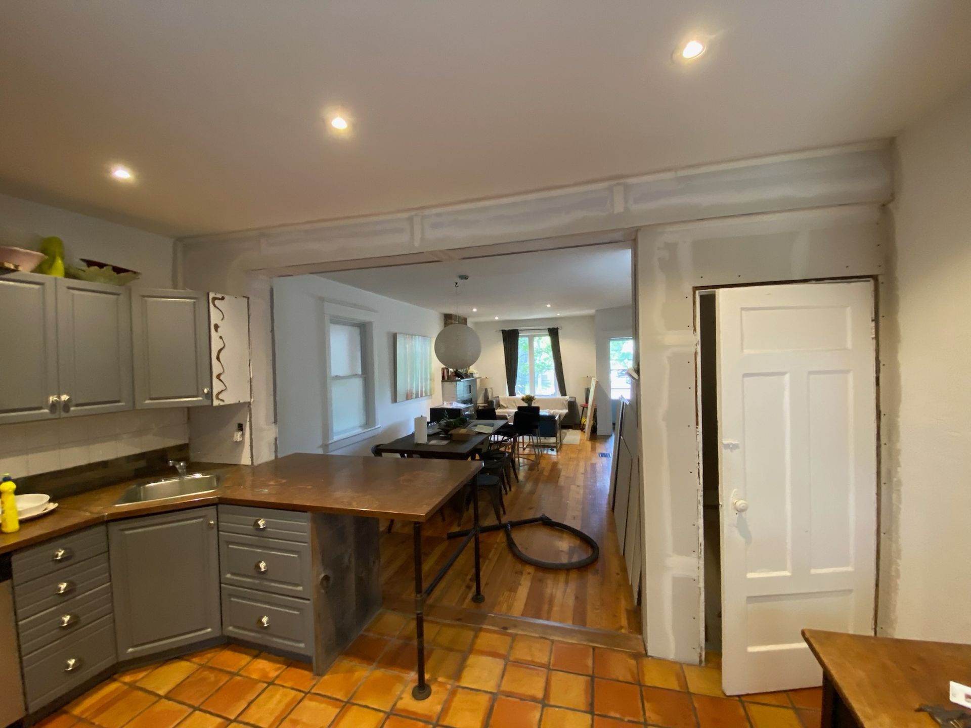 Kitchen renovation in progress: gray cabinets, open doorway to living area, drywall, and terra cotta floor.