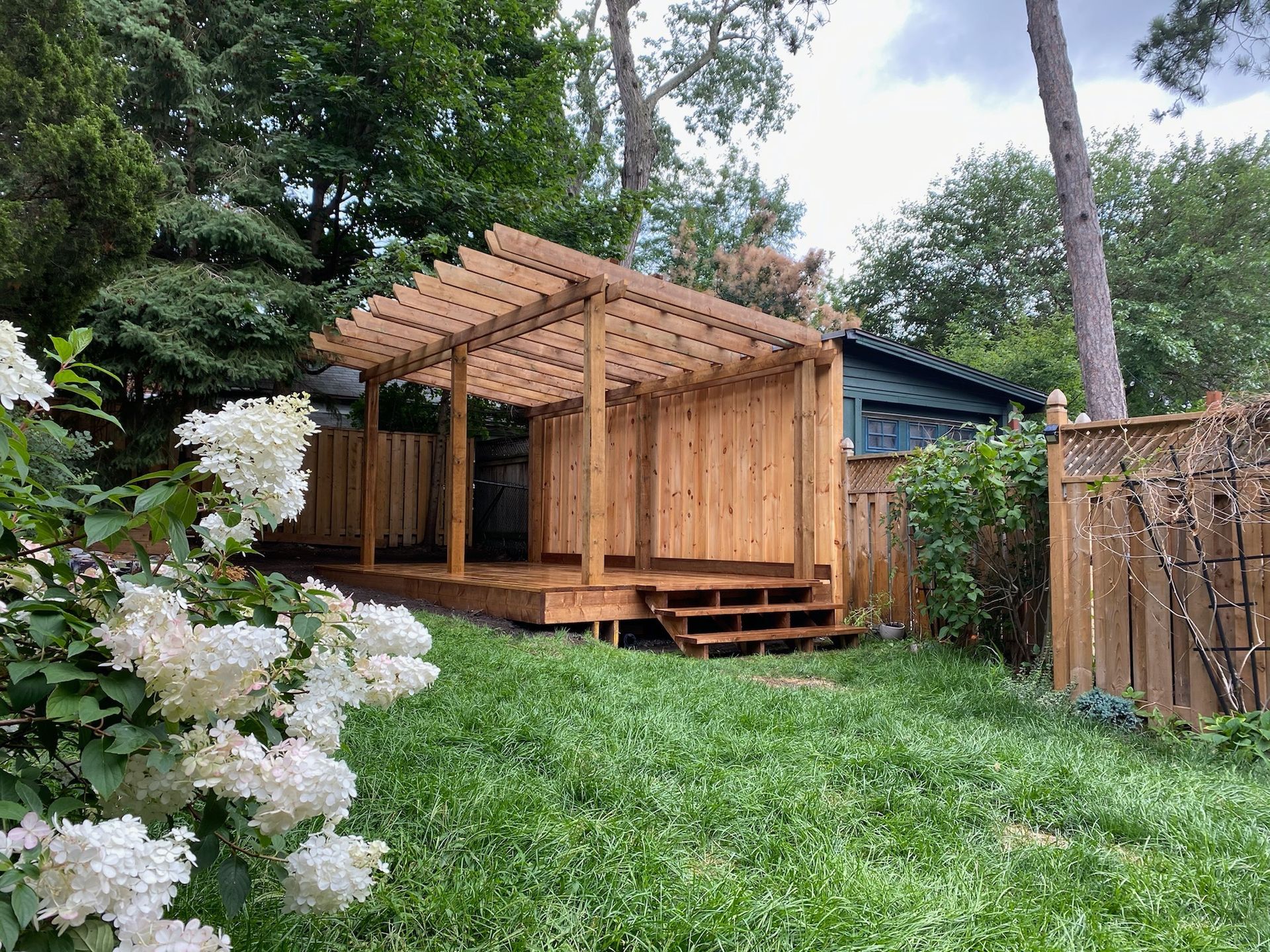 Wooden pergola structure on a raised deck with steps, surrounded by greenery and a fence.