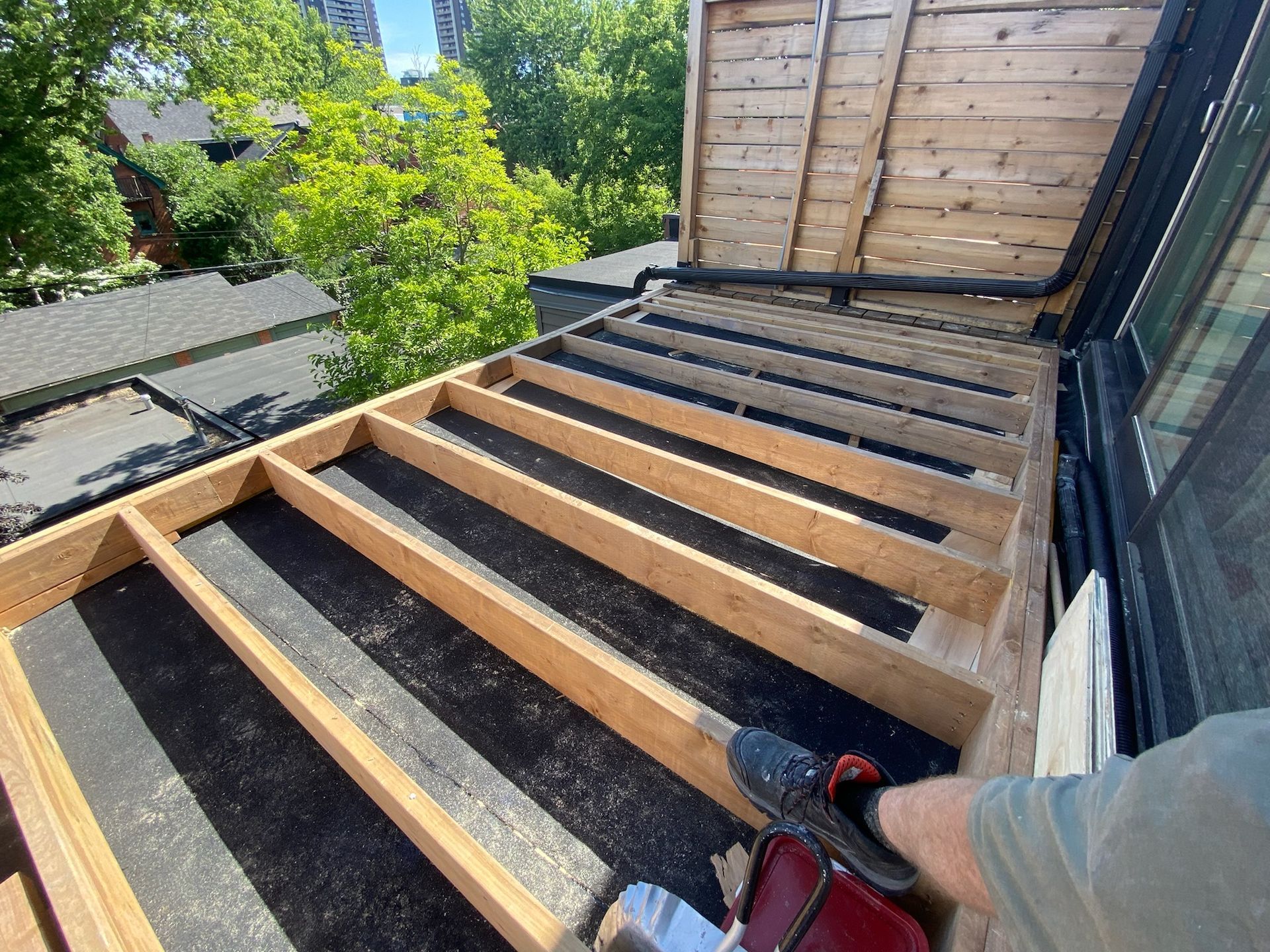 Construction of a wooden deck. A person uses a drill on wood beams and black underlayment.
