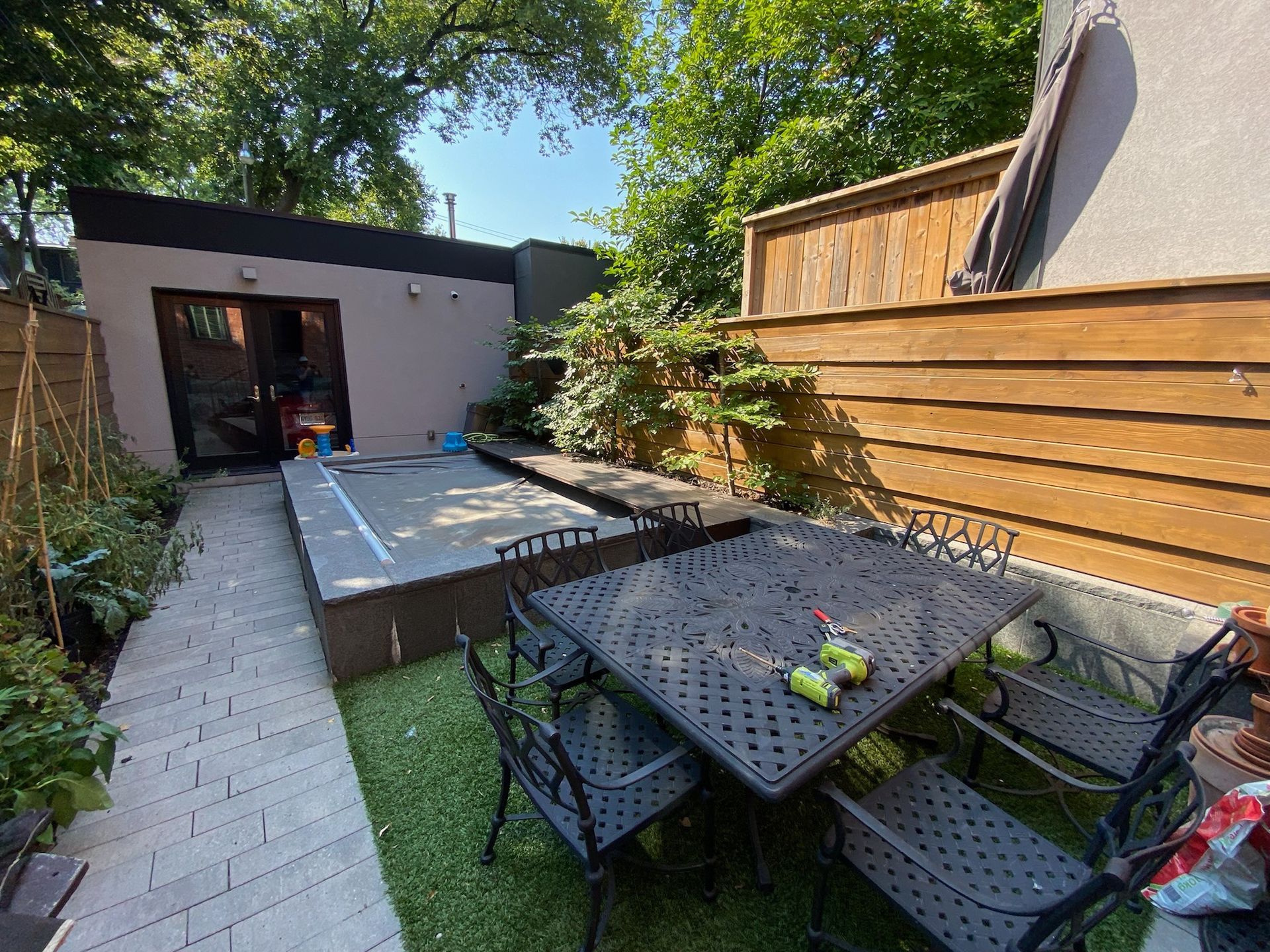 Backyard patio with table, chairs, hot tub, and modern building. Lush greenery.