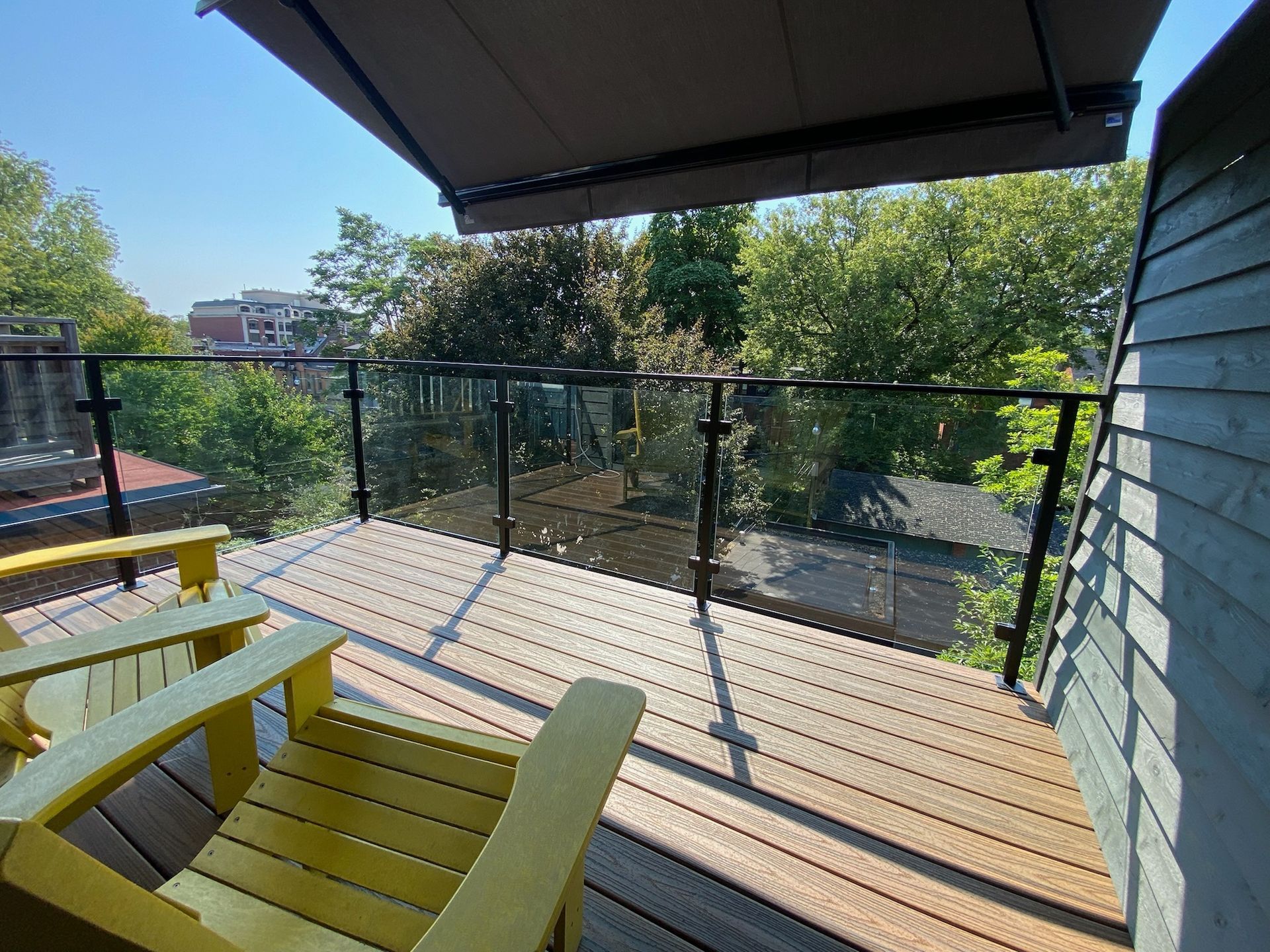 Two yellow Adirondack chairs on a wooden deck with glass railing, awning, and trees in the background.