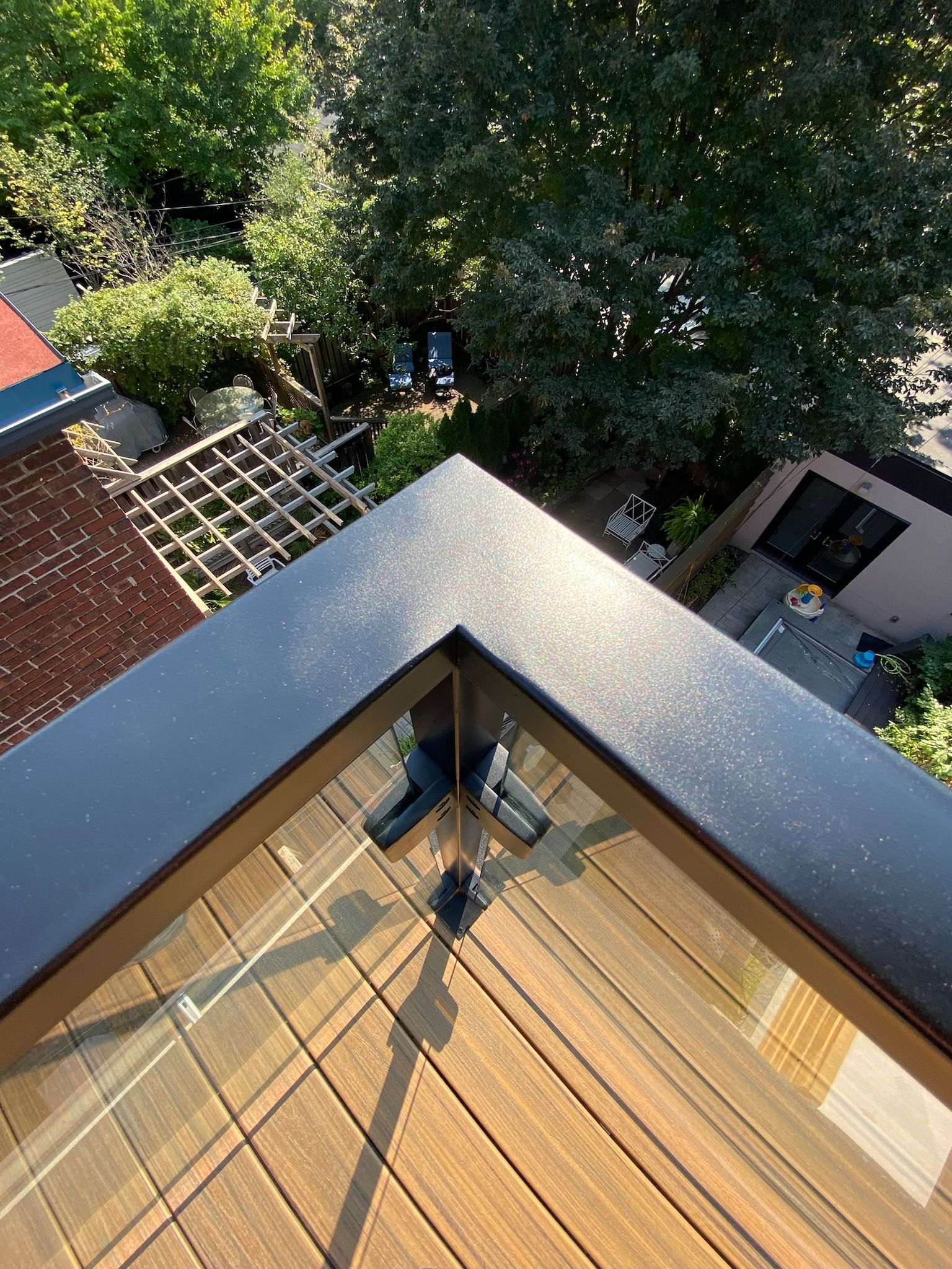 Overhead view of a deck corner with black railing, glass panels, and a brown wooden floor, overlooking greenery.