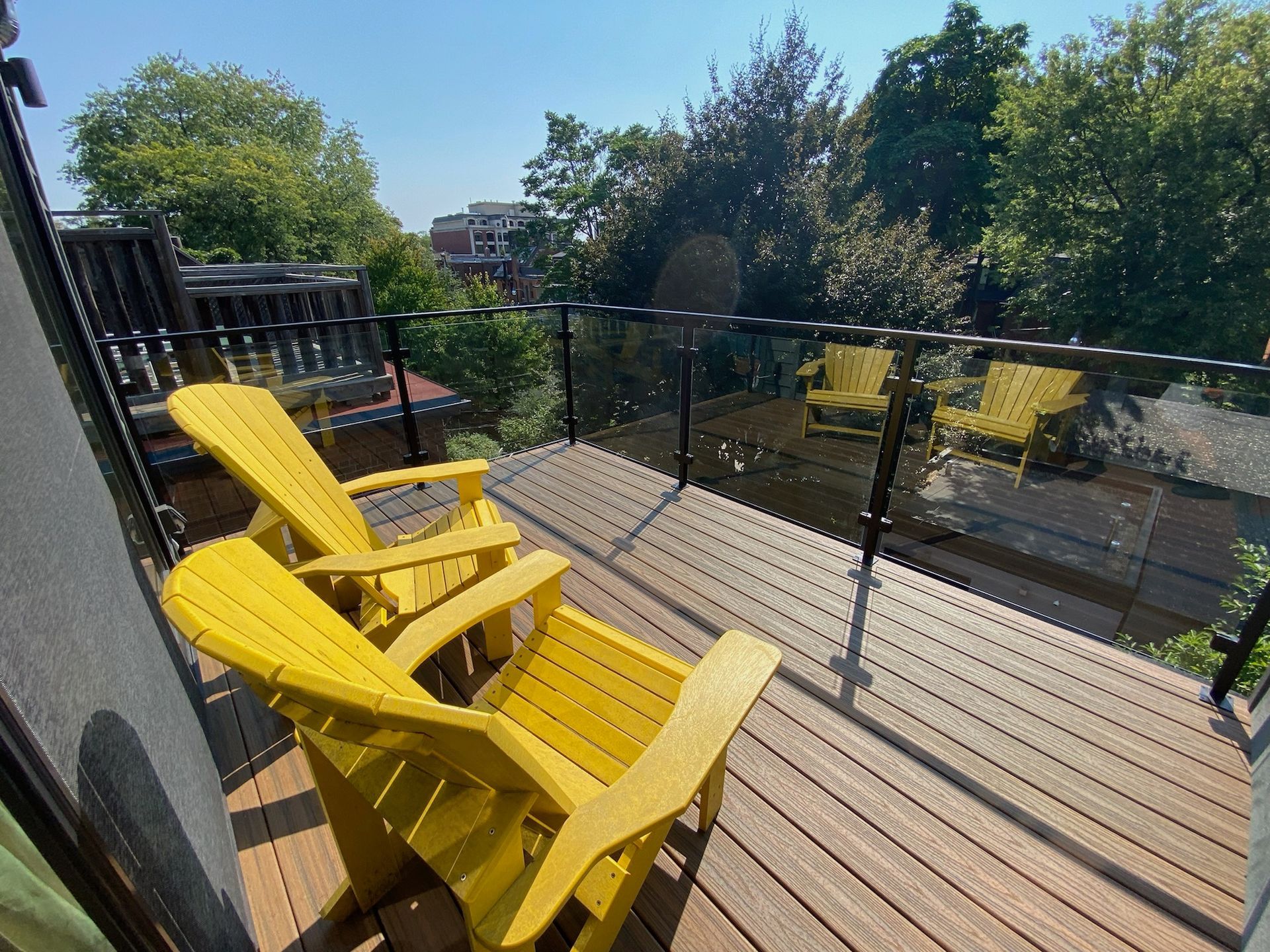 Two yellow Adirondack chairs on a wooden deck with glass railing, overlooking trees and other buildings.