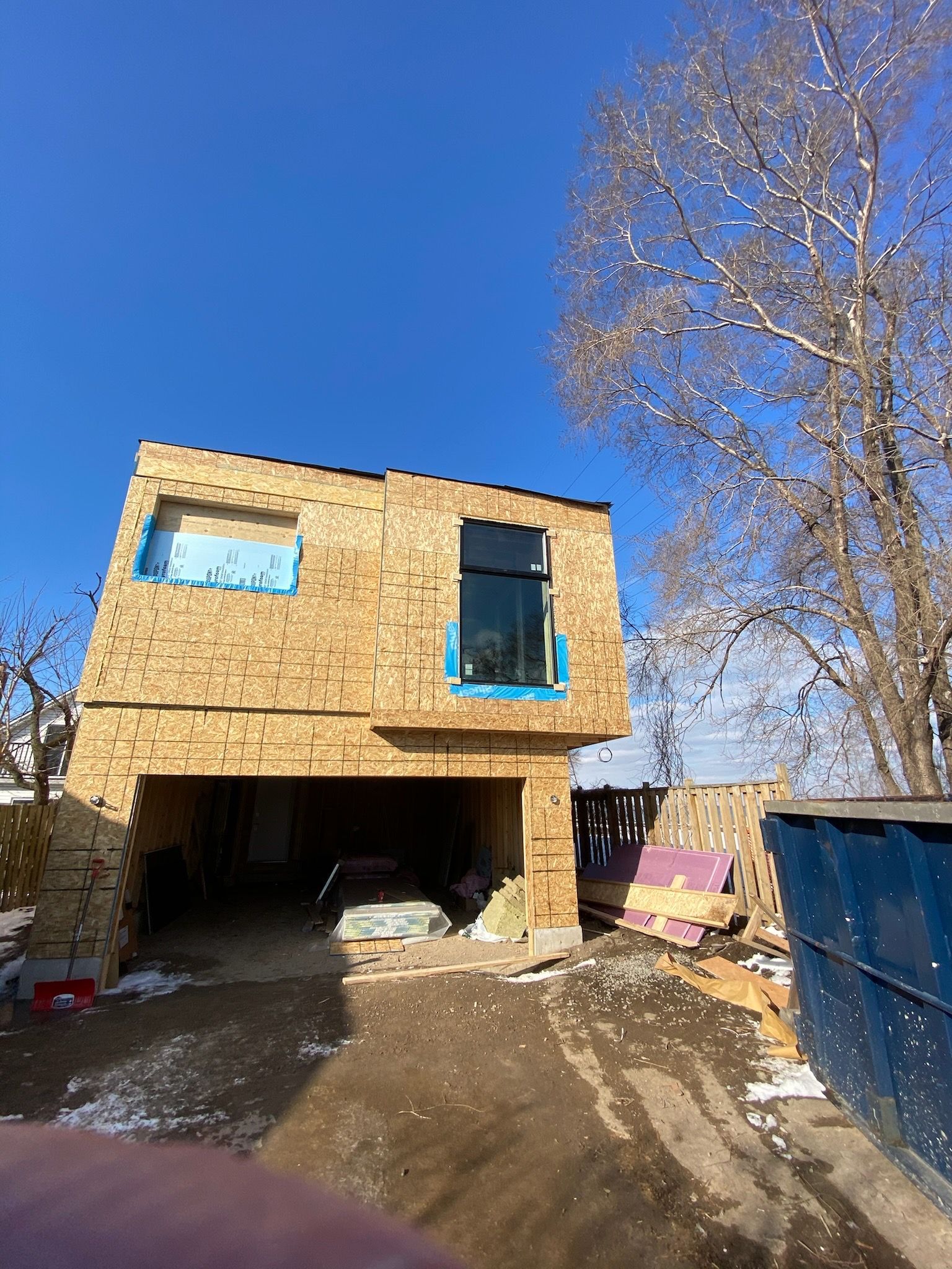 Garage under construction with OSB siding, a dark window, blue sky, and a bare tree.
