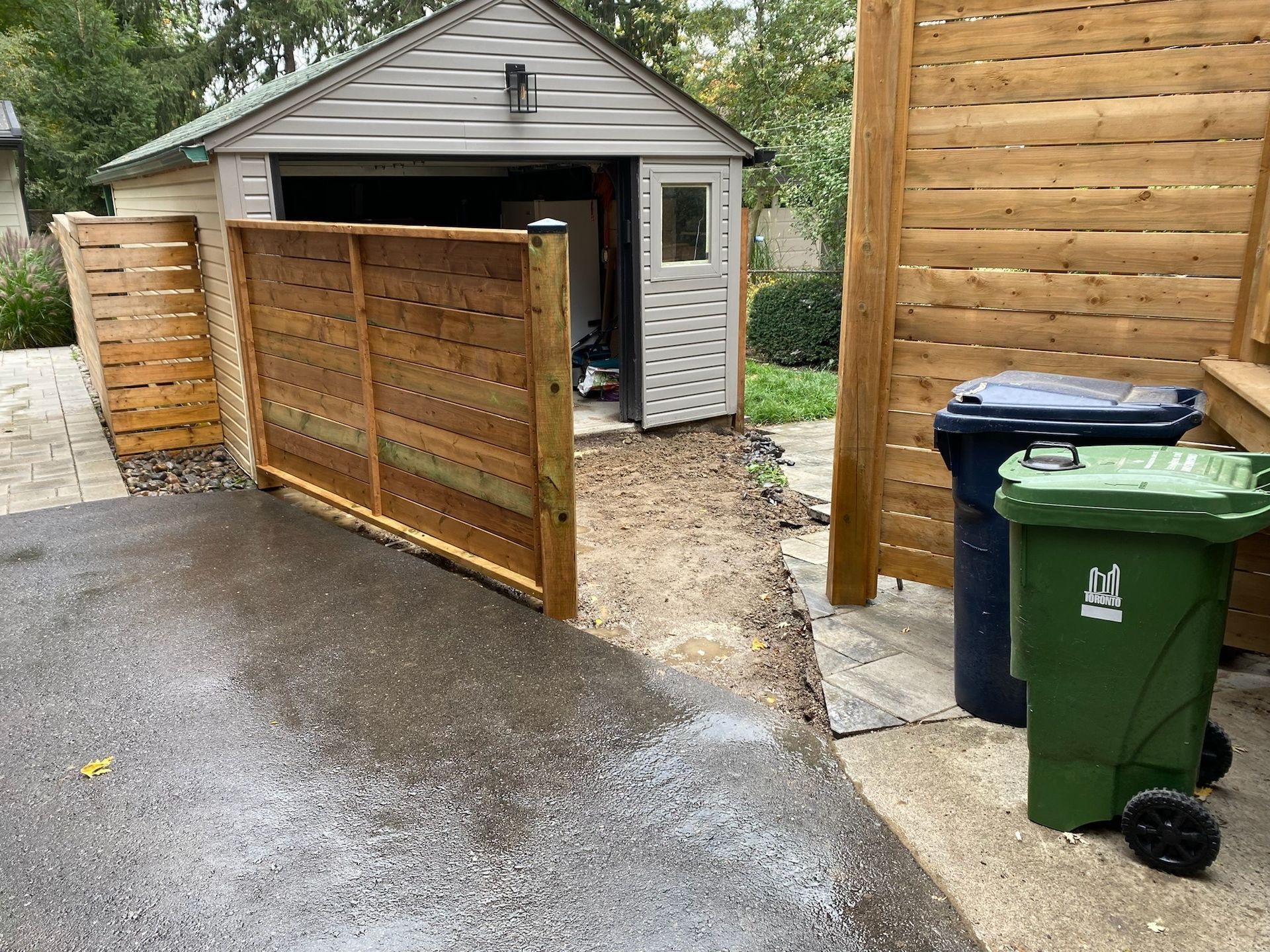 A driveway leads to a garage. Wooden fences border the driveway. Two trash bins stand nearby.