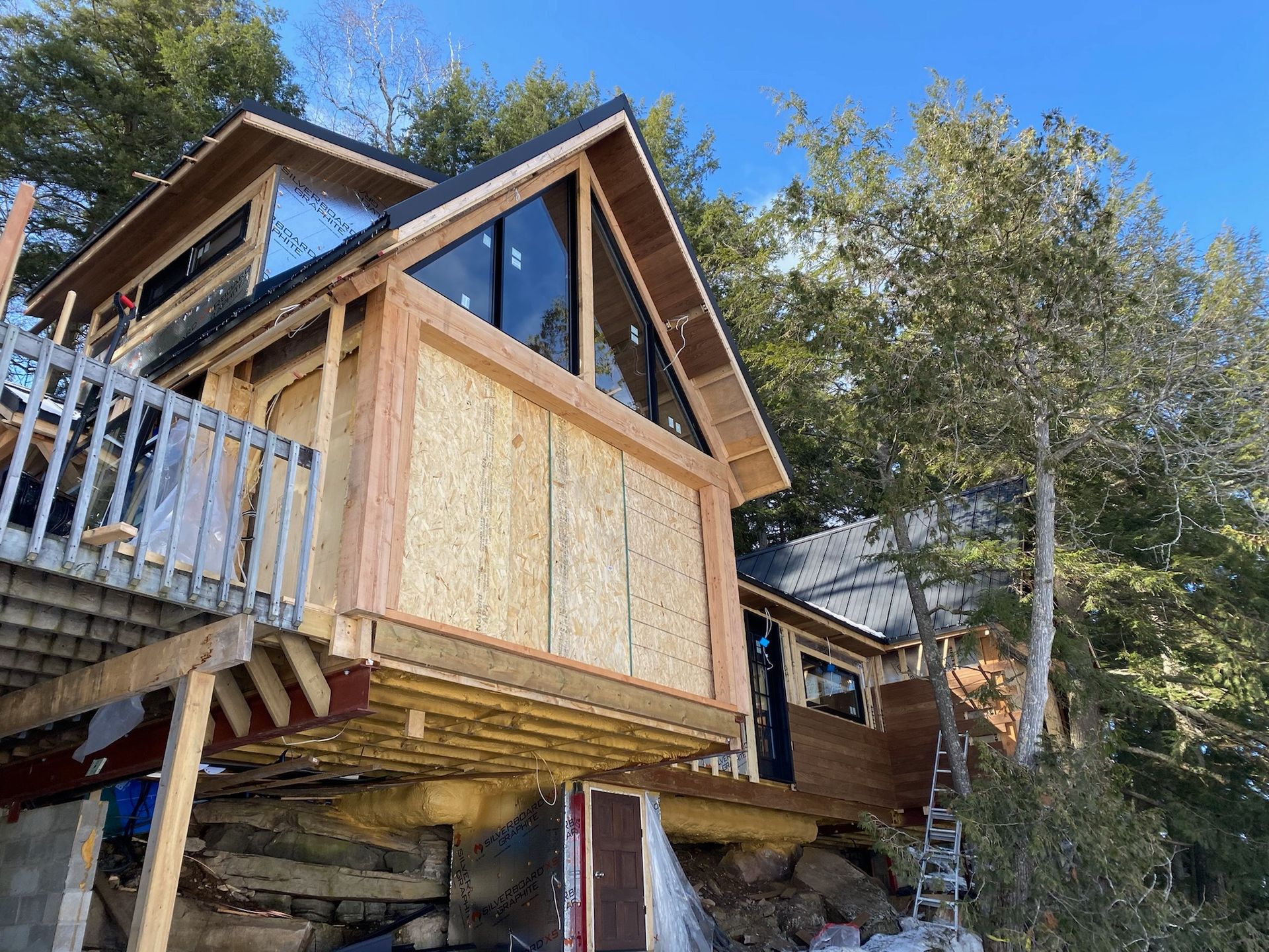 Cabin under construction on a hillside with large windows and a wooden deck.