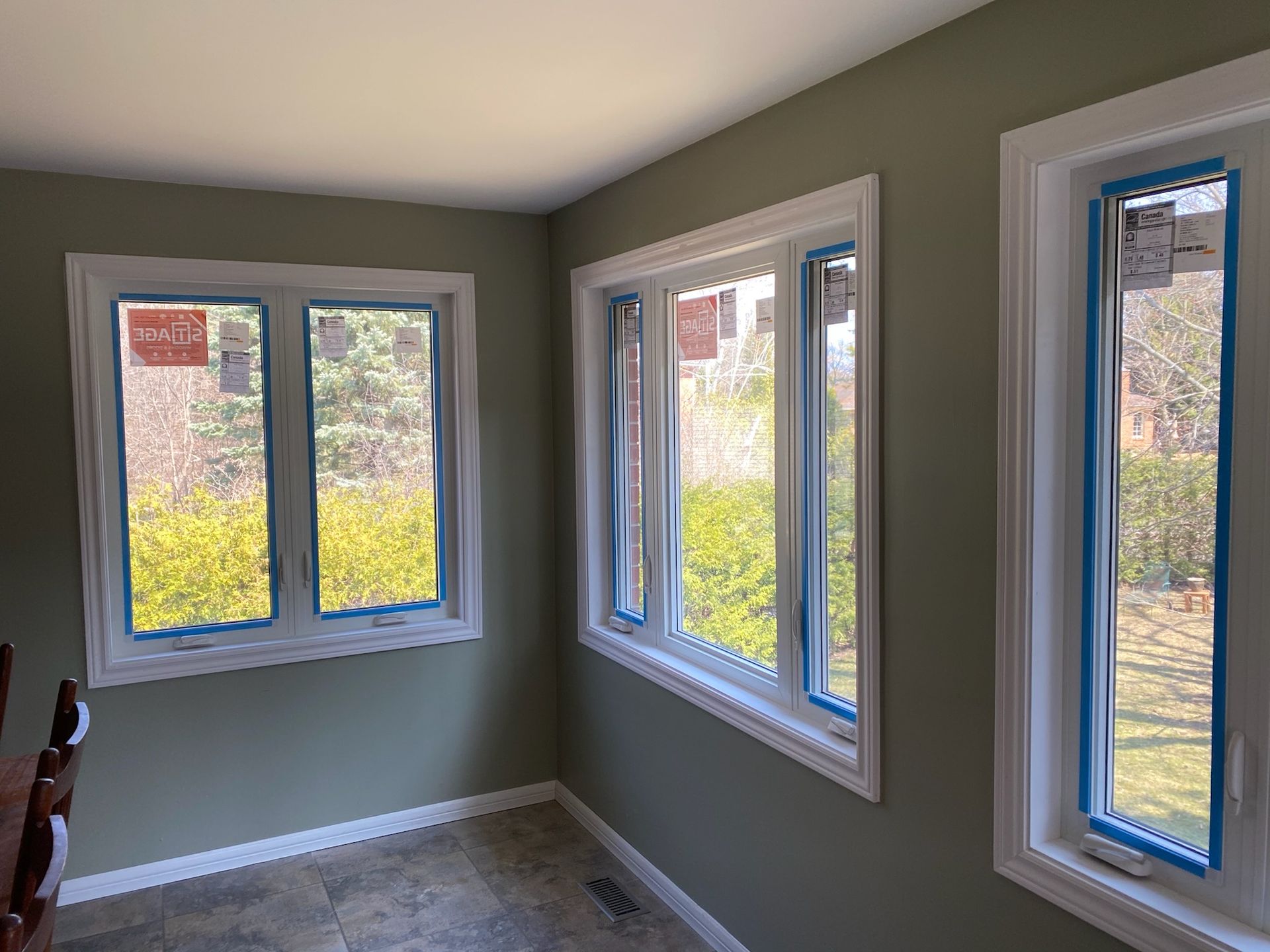 Three white-framed windows on a green wall. New construction with protective blue tape, view of trees outside.
