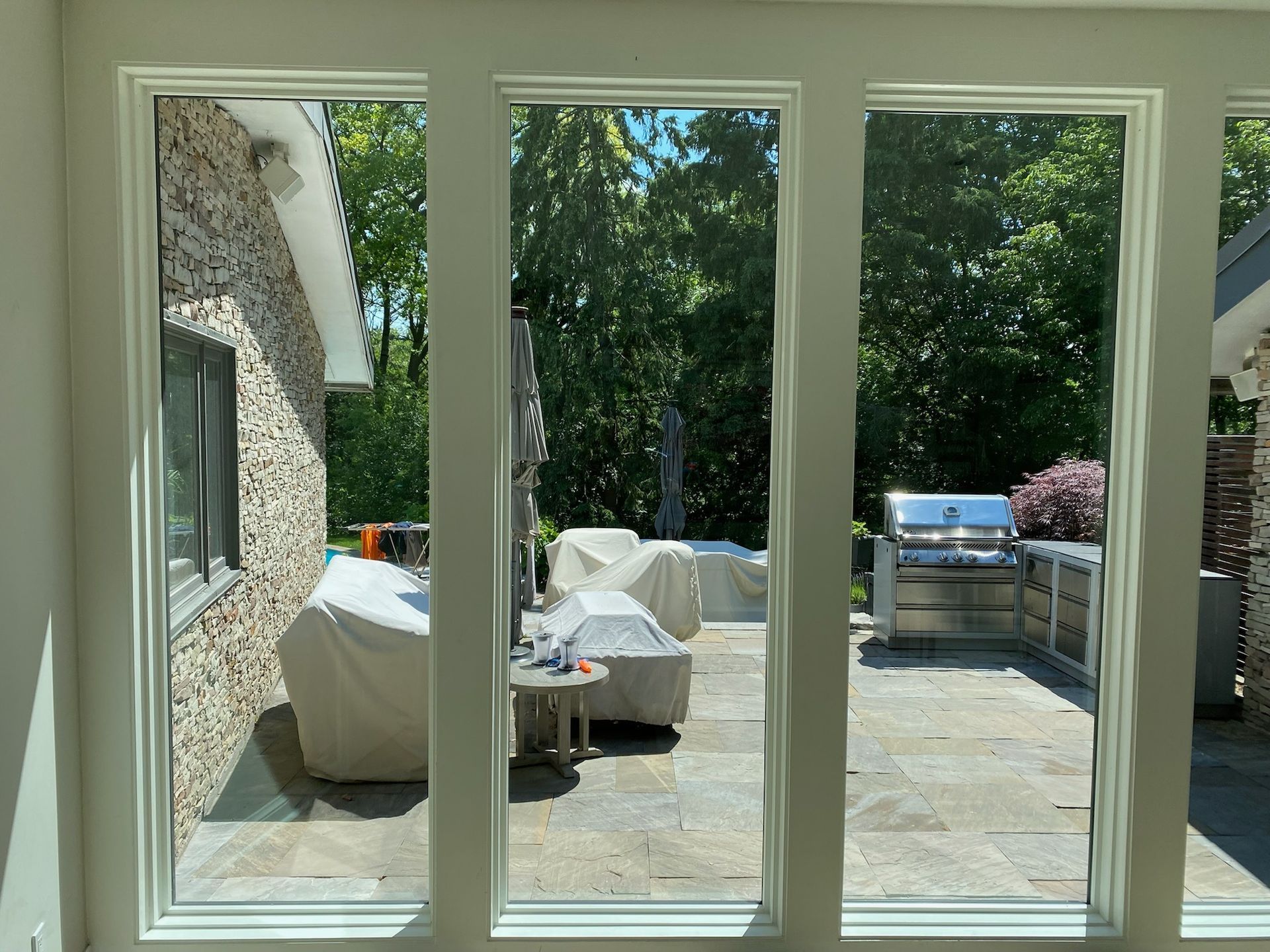 Three tall, white-framed windows offer a view of a stone patio with covered furniture, a grill, and lush trees.