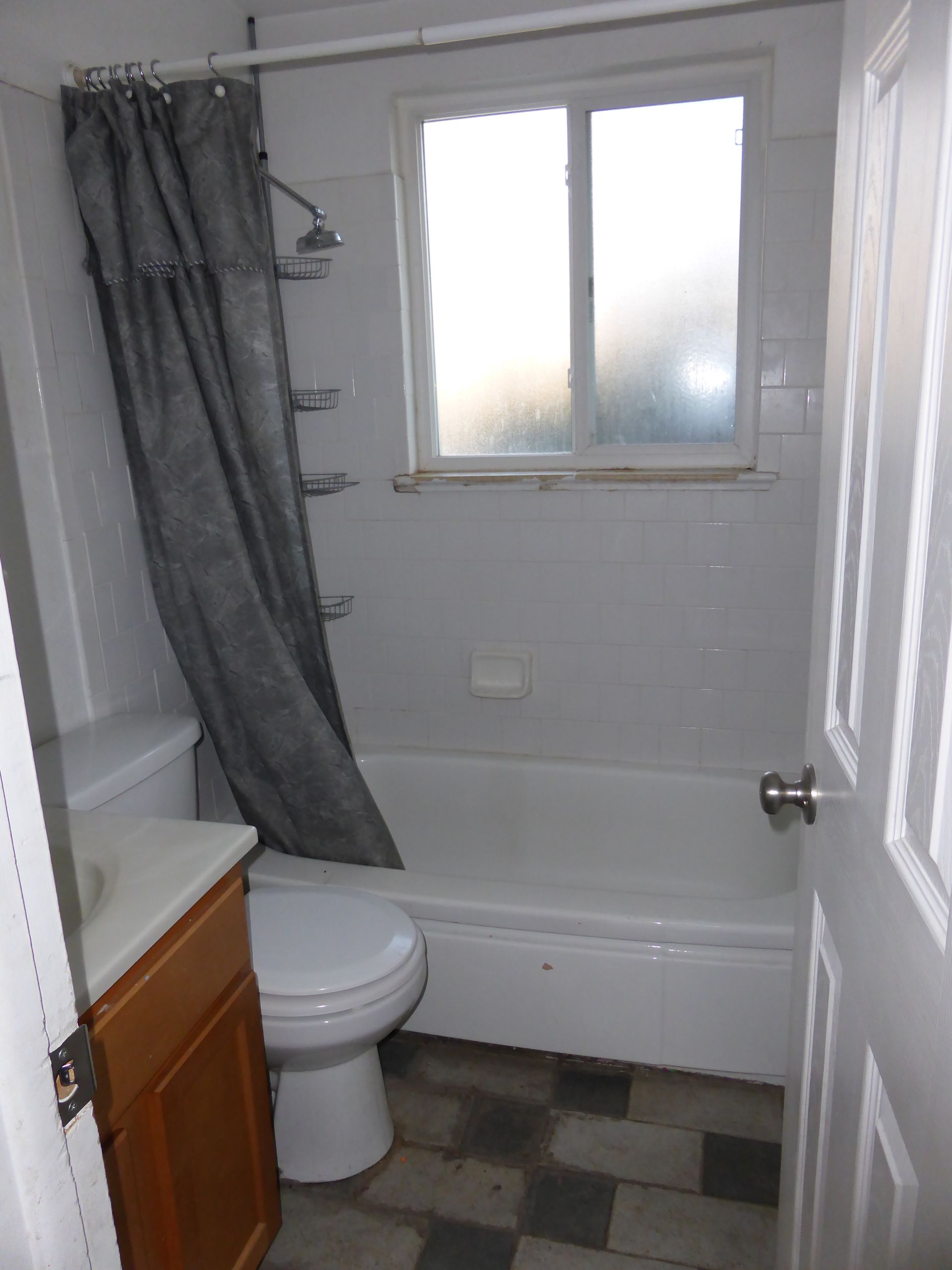 Small bathroom with a toilet, sink, bathtub, frosted window, and gray shower curtain