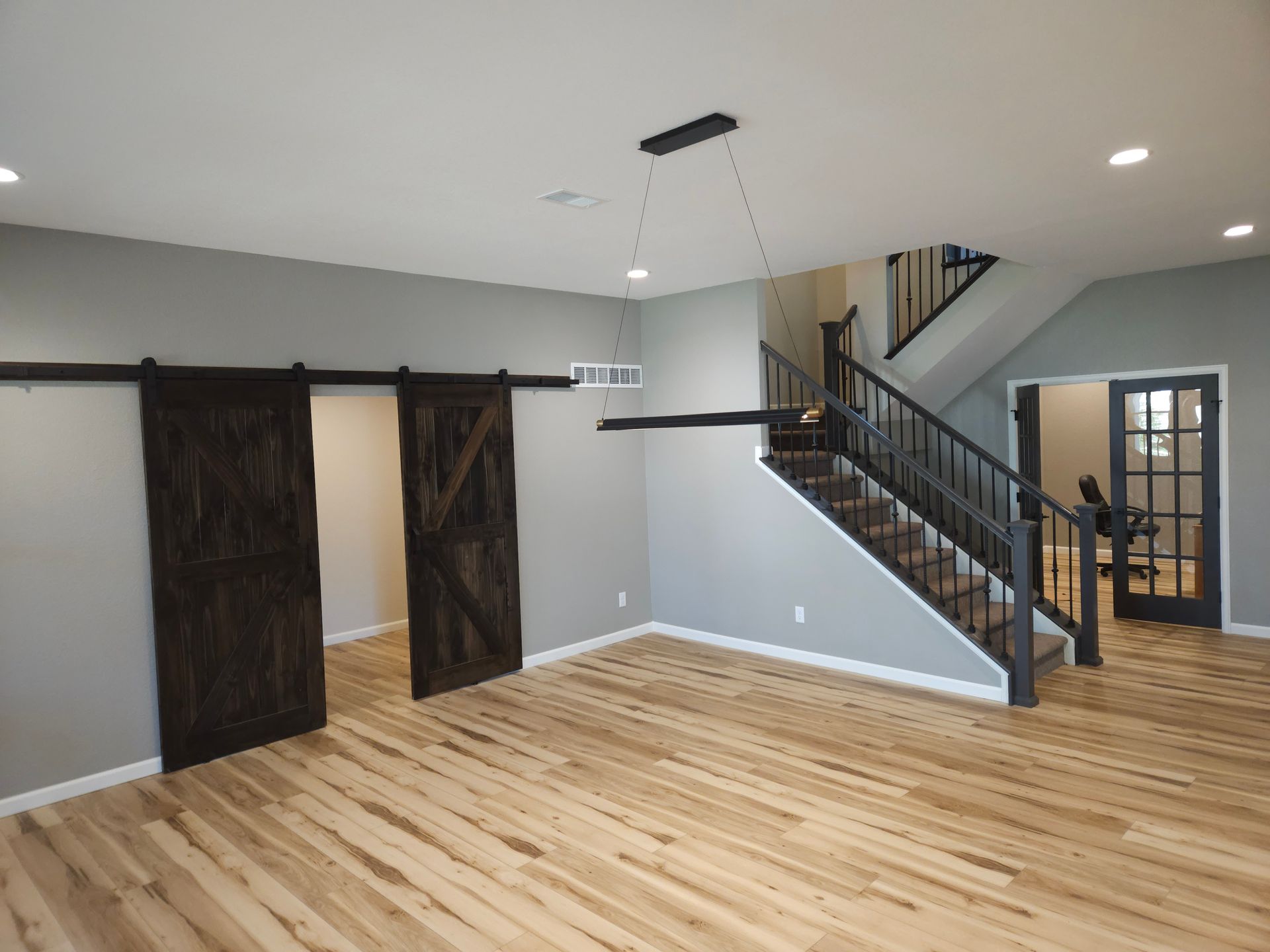 Empty room with wood floors, dark sliding barn doors, staircase, and a ceiling light over a kitchen pass-through