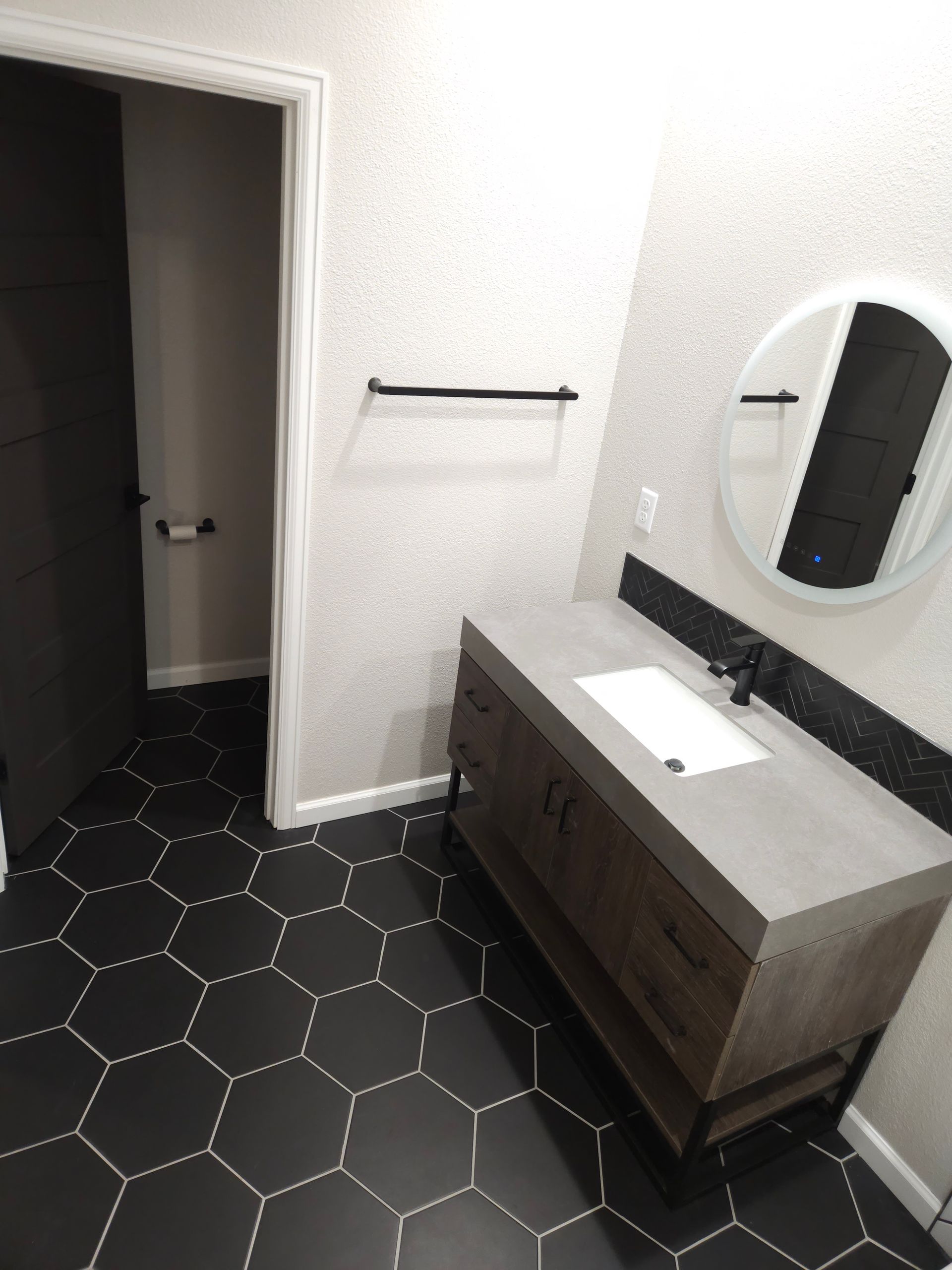 Small bathroom with black hex tile floor, gray vanity, oval mirror, and towel bar by an open doorway.