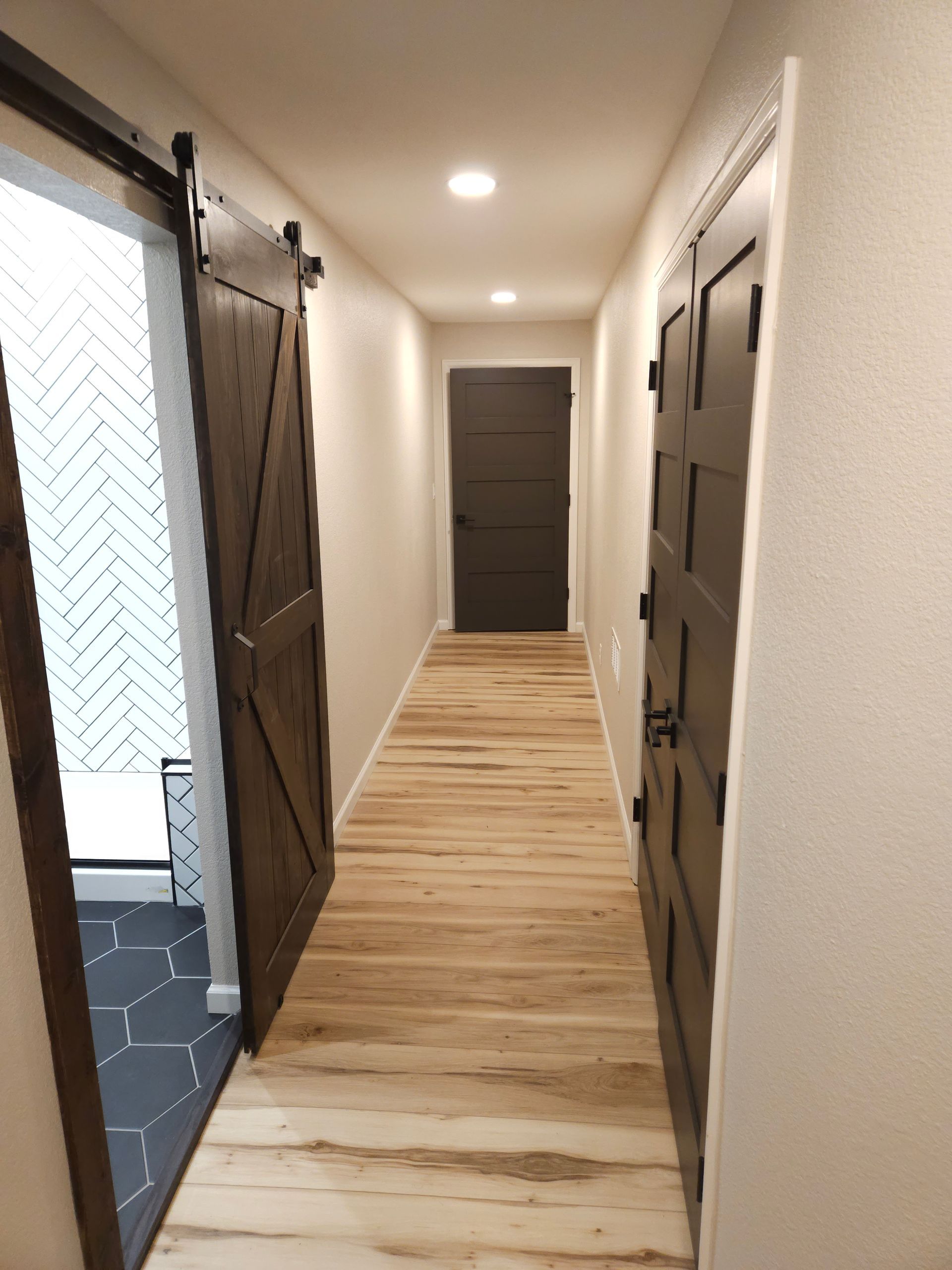 Narrow hallway with light wood floors, white walls, open dark doors, and a windowed room on the left.