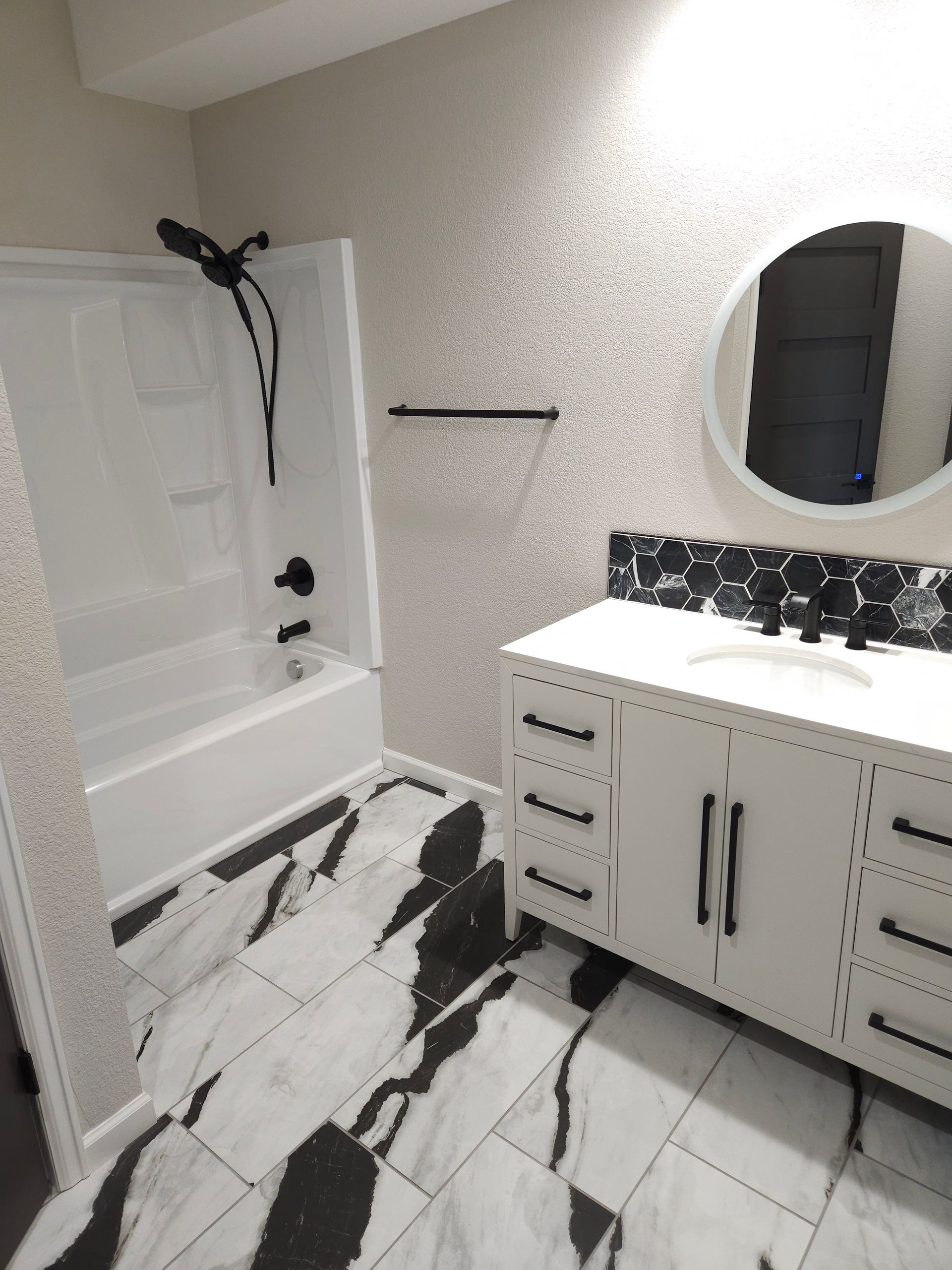 Modern bathroom with white vanity, round mirror, tub, and black-and-white marble floor tiles