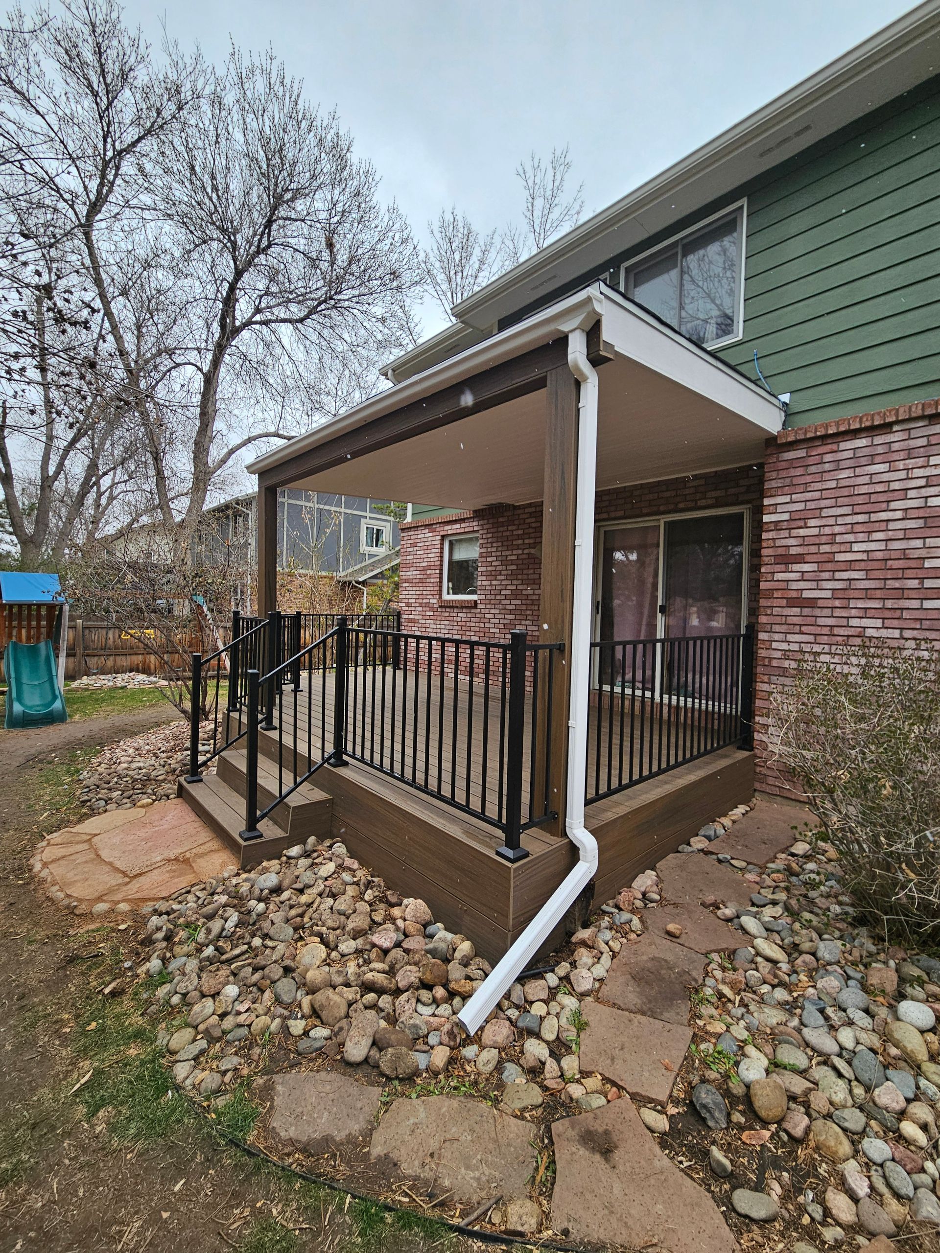 Small covered porch with black railing and white downspout beside a green house, on a rocky yard.