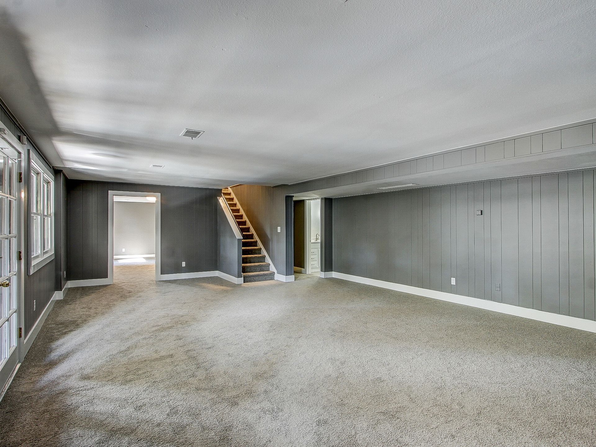Empty carpeted room with gray paneled walls and a staircase at the back-left.