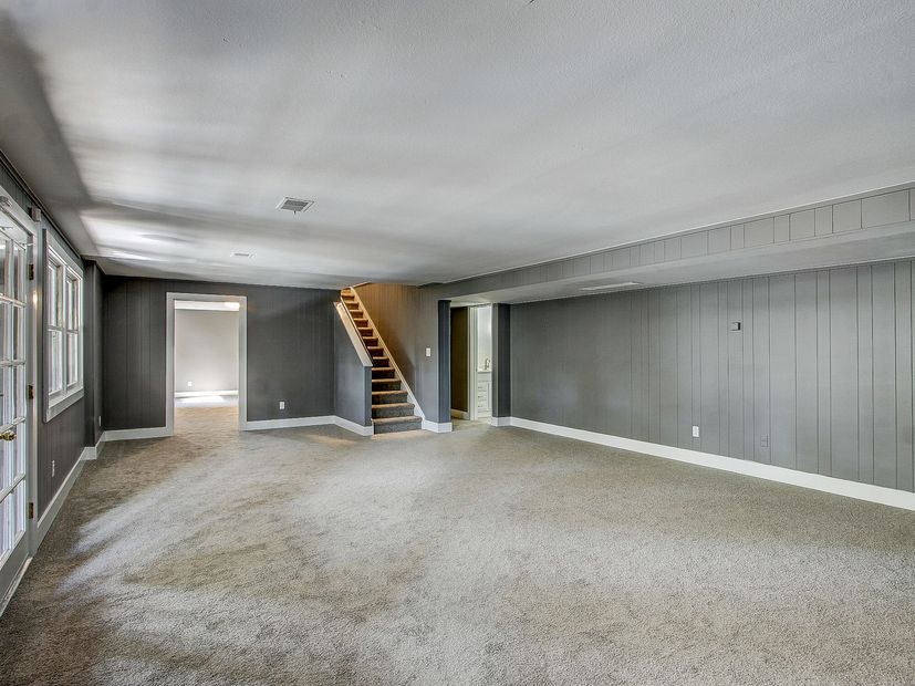 Empty carpeted living room with gray walls, a doorway, and a wooden staircase.