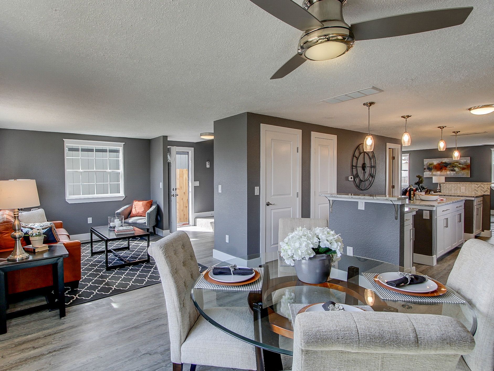 Open-concept living room and kitchen with gray walls, ceiling fan, and glass dining table in the foreground