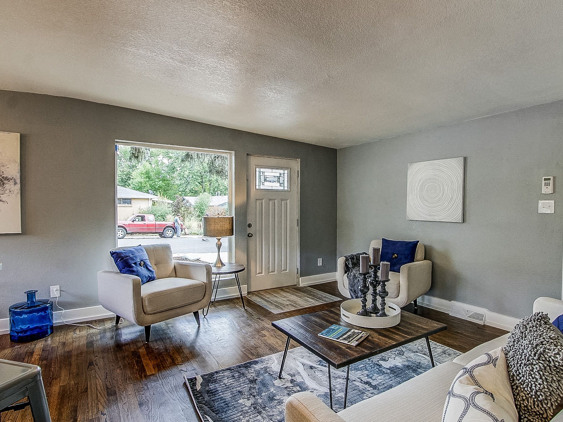 Bright living room with gray walls, beige chair, coffee table, rug, and open front door to a sunny yard.