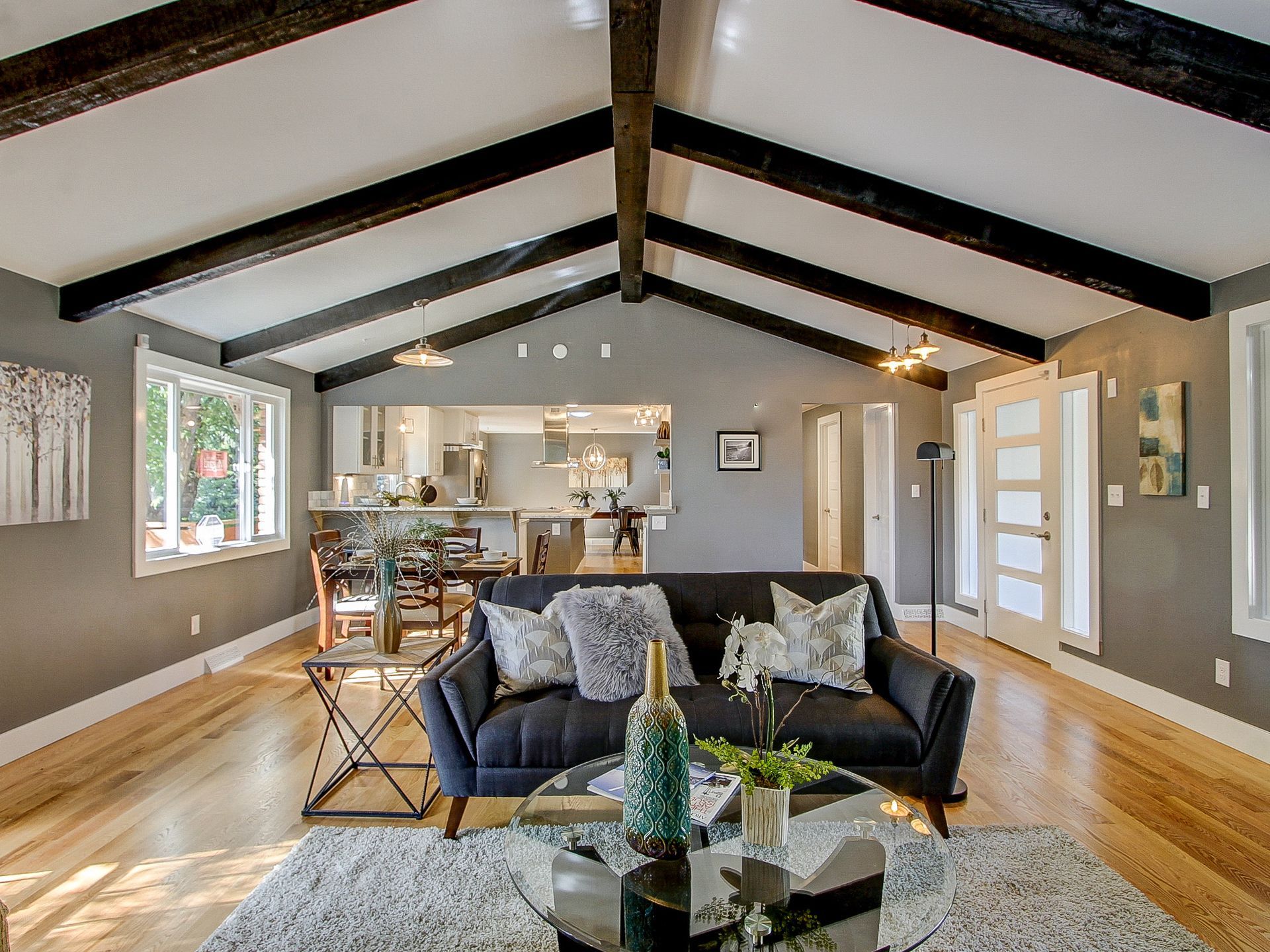 Bright open living room with vaulted ceiling, black beams, gray sofa, and hardwood floors leading to a kitchen.