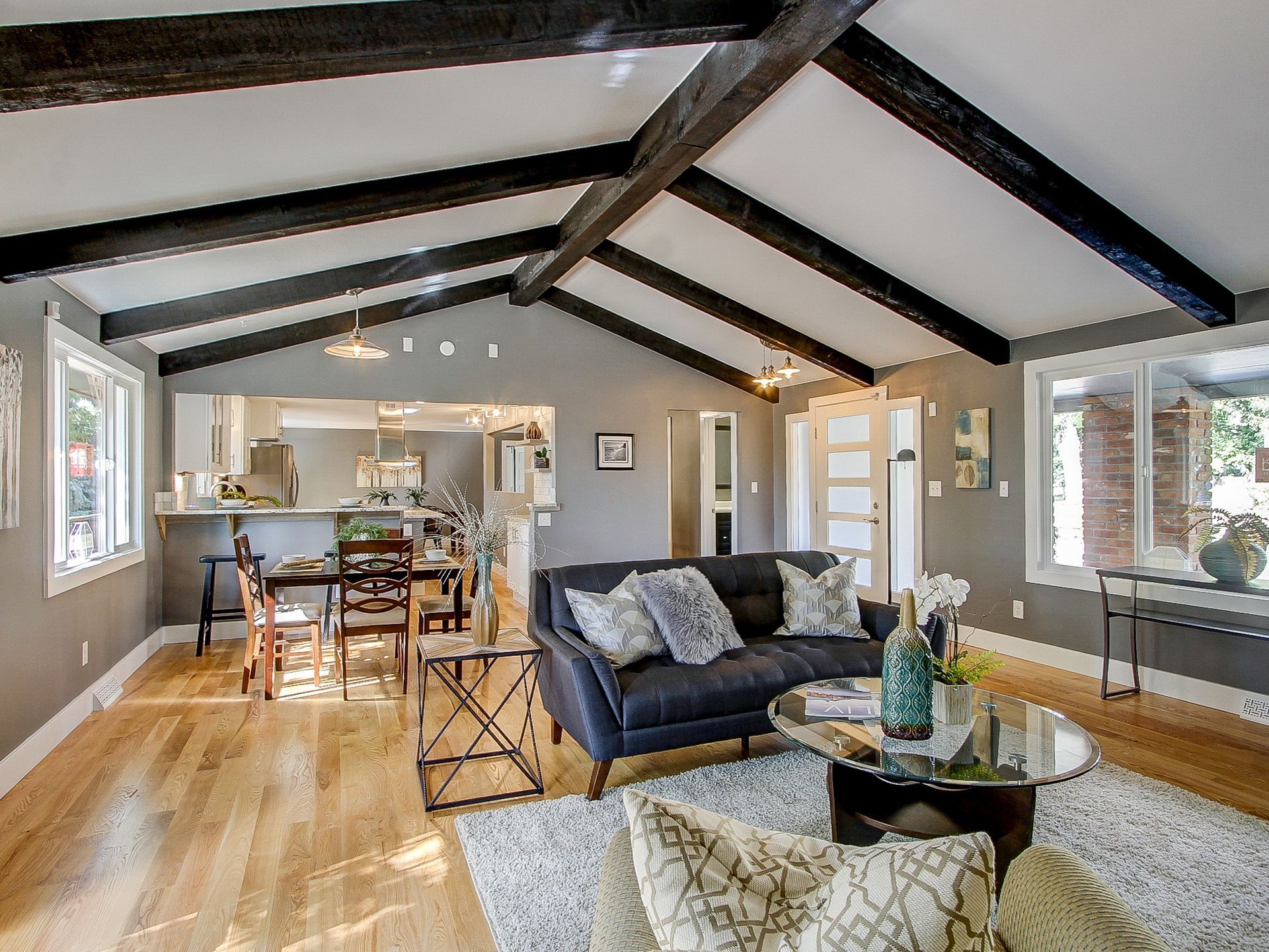 Bright open living room with vaulted ceiling, dark beams, gray sofa, and dining area beyond