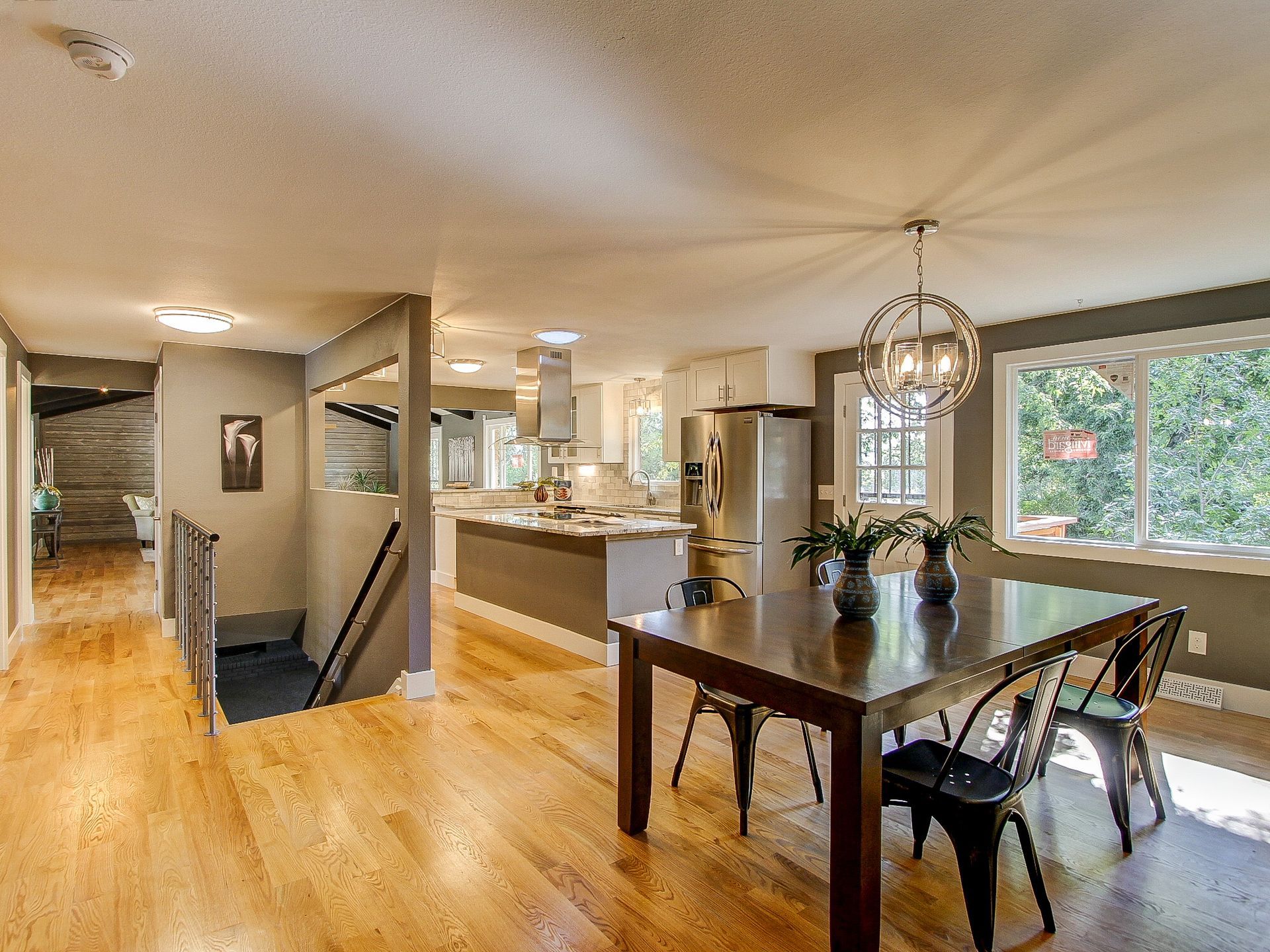 Open-plan home interior with kitchen, dining table, hardwood floors, and large windows overlooking greenery