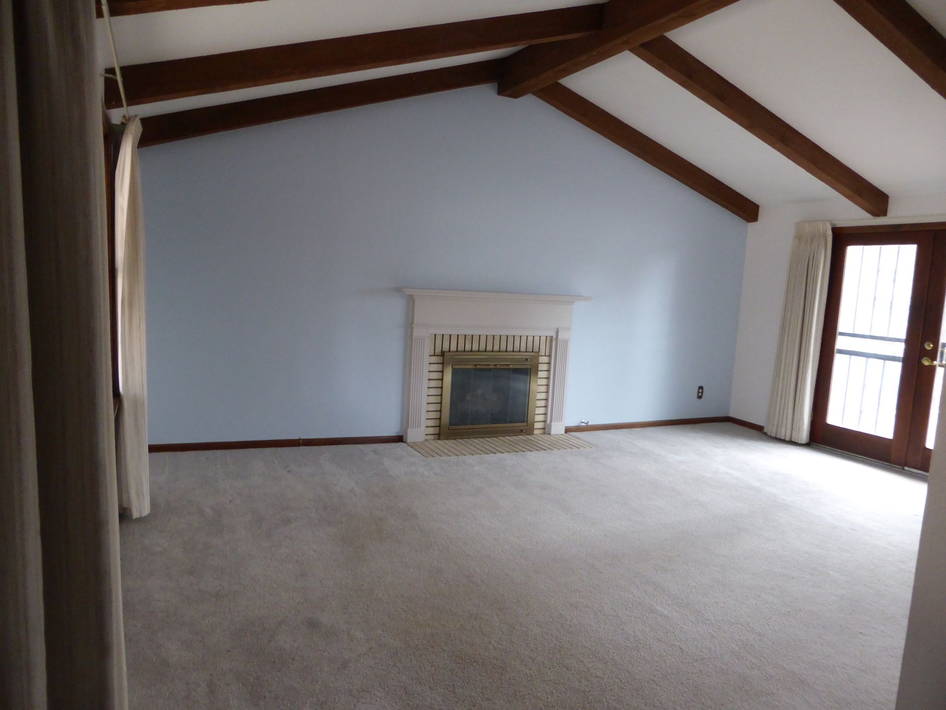 Empty living room with vaulted wood-beam ceiling, fireplace, carpeted floor, and large windows.