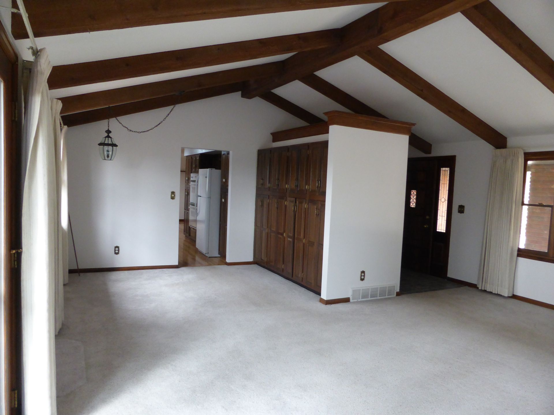 Empty carpeted room with exposed wooden beams, white walls, closets, and a window.