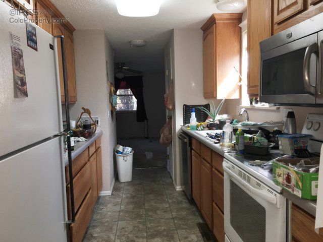 Narrow kitchen with wooden cabinets, white appliances, and cluttered countertops leading to a dark hallway.