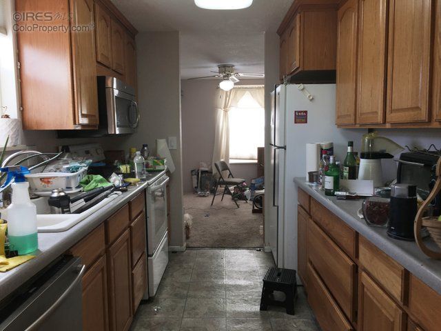 Narrow kitchen with wood cabinets, white fridge, cluttered countertops, and a view into a carpeted dining area.