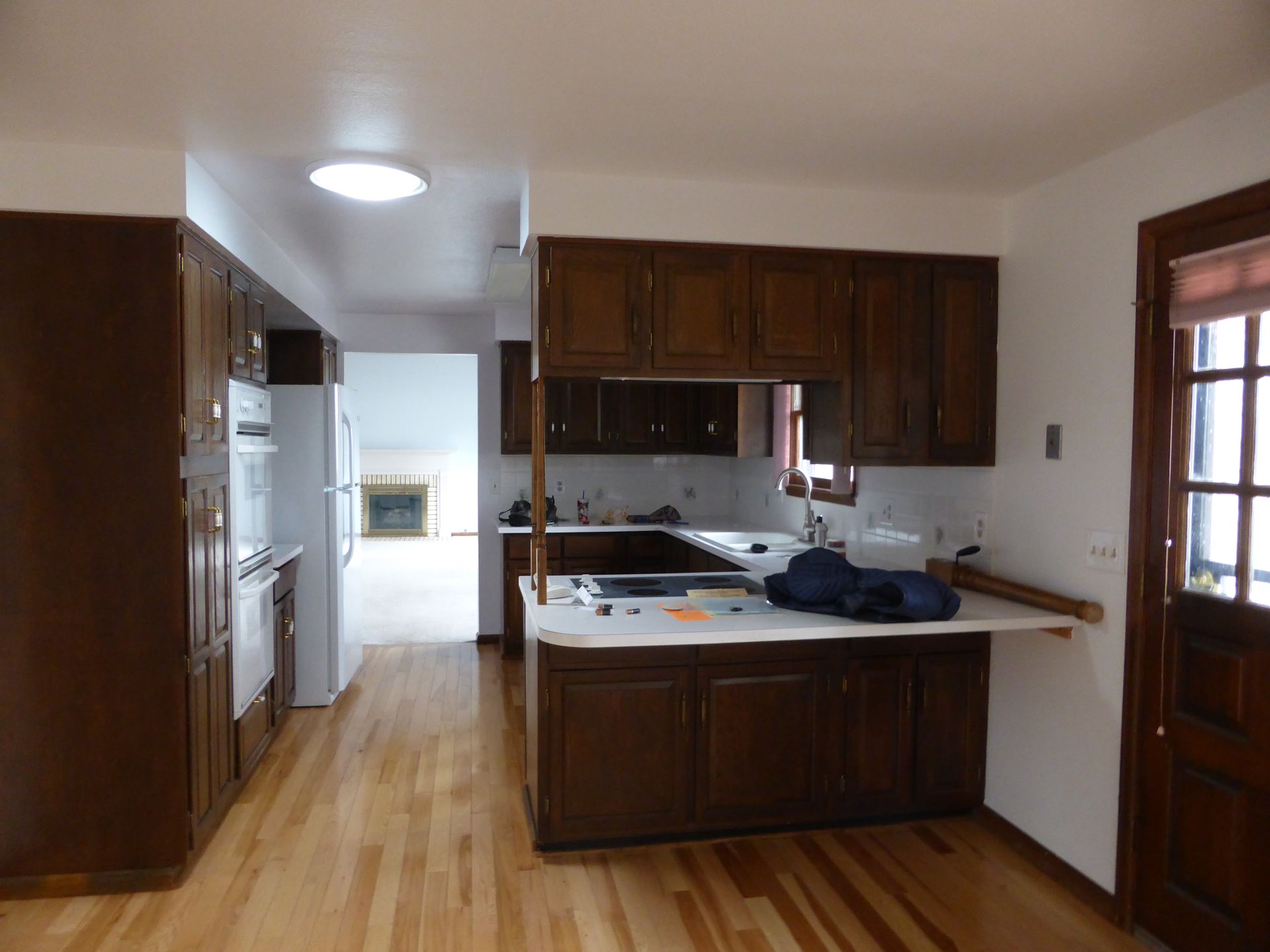 Empty kitchen with dark wood cabinets, white walls, and hardwood floors, viewed toward a bright hallway.