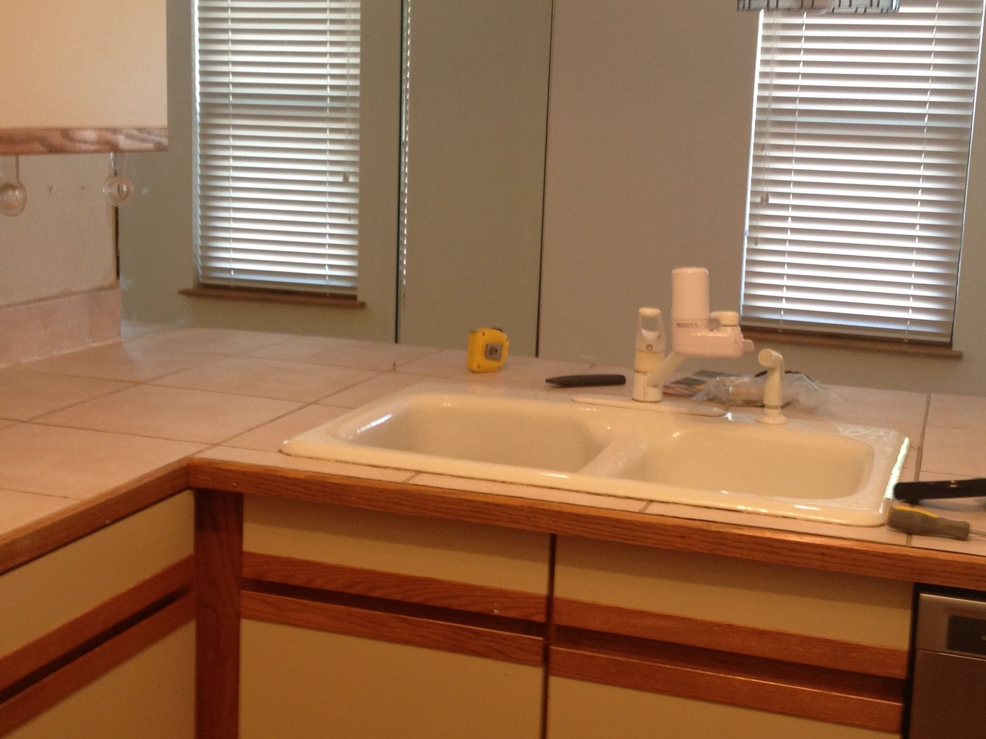 Bathroom sink and countertop with beige tiles, white fixtures, and two windows with blinds.