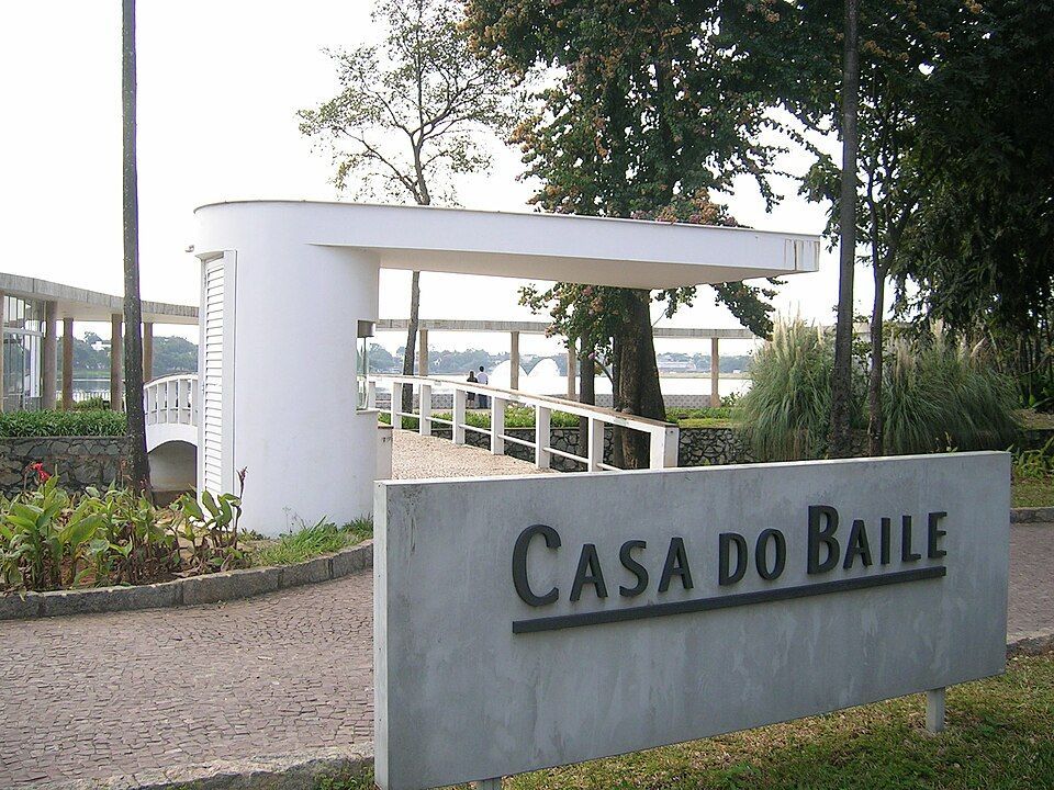 Placa de entrada da Casa do Baile, com edifício branco e árvores ao fundo.