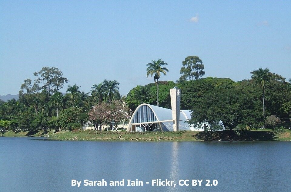 Monumento em arco branco e chaminé alta à beira de um lago, emoldurados por árvores sob um céu azul.