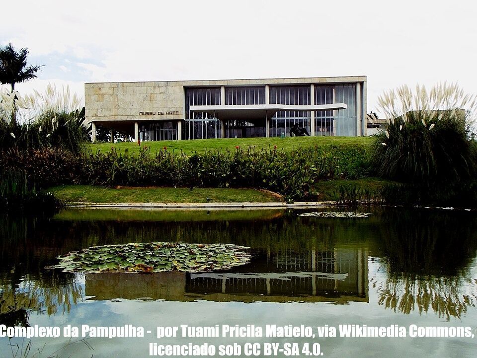 Prédio moderno atrás de um lago com nenúfares e vegetação densa sob um céu nublado.
