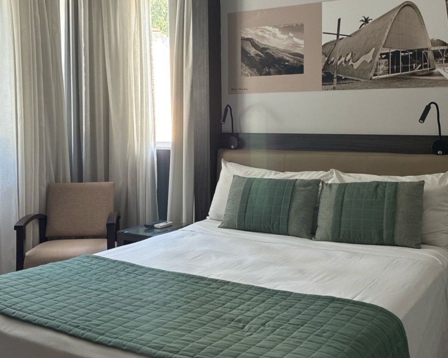 Cama arrumada com colcha verde em um quarto moderno, ao lado de uma cadeira e janela com cortinas transparentes.