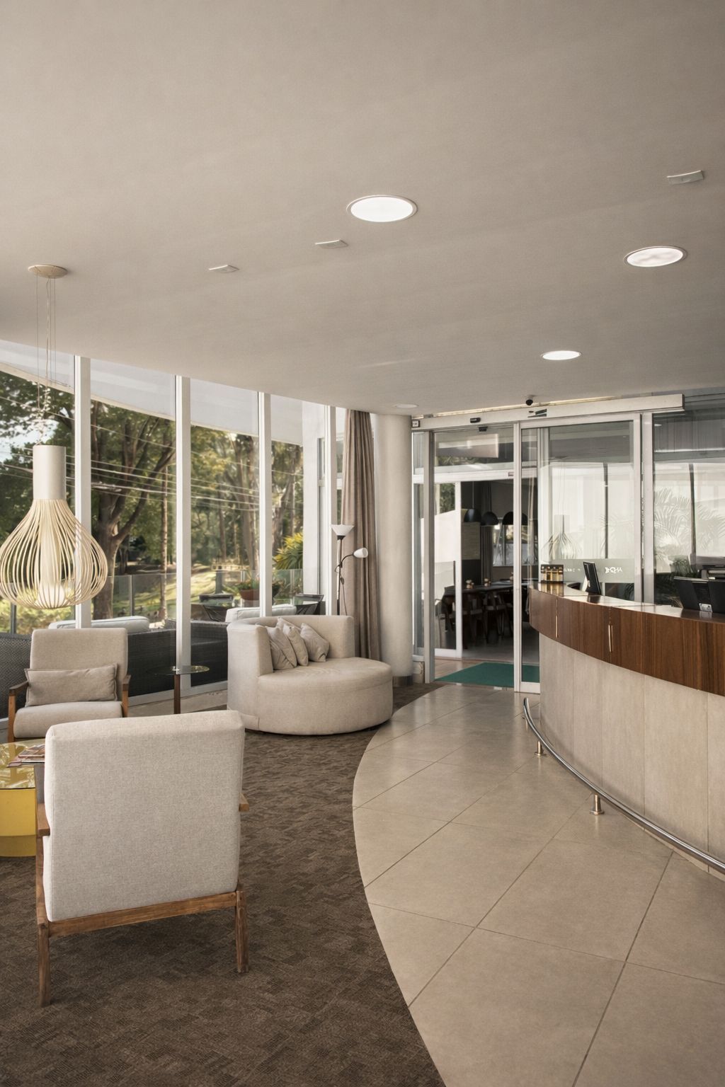 Hall de entrada moderno do hotel com balcão de recepção curvo, poltronas e grandes janelas com vista para a vegetação.