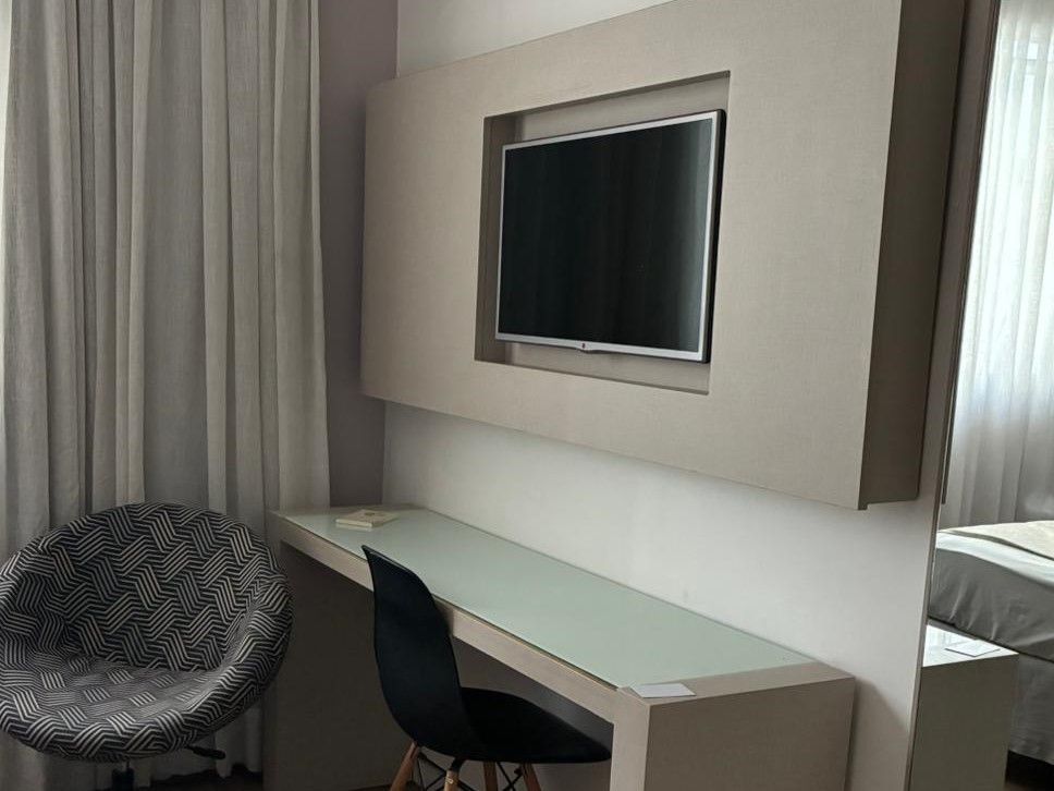 Área de escritório moderna em quarto de hotel com TV fixada na parede, cadeira preta, cadeira de vime e cortinas.